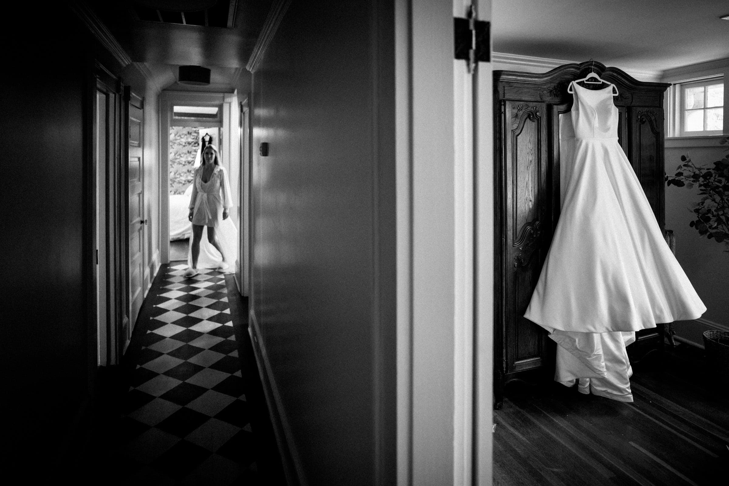 In a dimly lit hallway at Pinehurst Resort, a black and white photo captures a bridal gown hanging on a wardrobe door. A person in a robe walks across the checkered floor in the background as sunlight filters through the window above, creating an atmosphere reminiscent of a quaint village chapel.