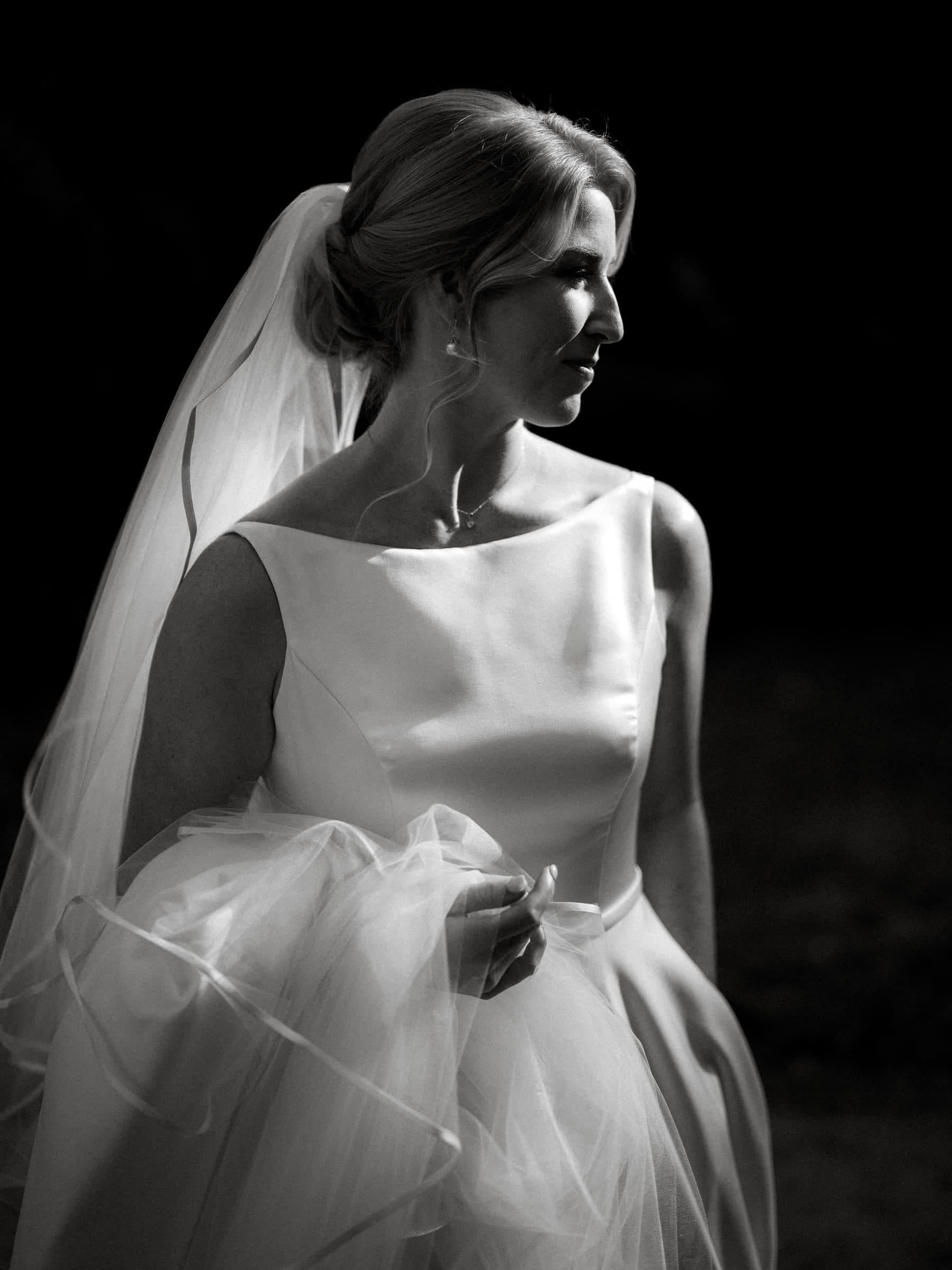 A bride in a white wedding dress stands gracefully outside the village chapel, holding her veil and skirt. Her hair is elegantly styled as she gazes thoughtfully to the side. The black and white setting at Pinehurst Resort casts a timeless, classic feel.