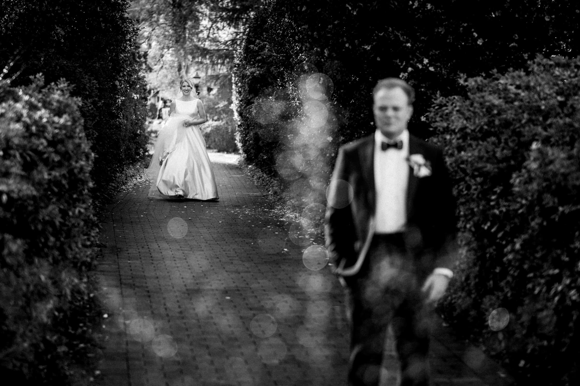 A black and white photo captures a bride in a flowing dress walking along a brick pathway lined with bushes at Pinehurst Resort. In the foreground, a groom in formal attire stands with his back facing her, evoking anticipation and emotion reminiscent of a village chapel moment.