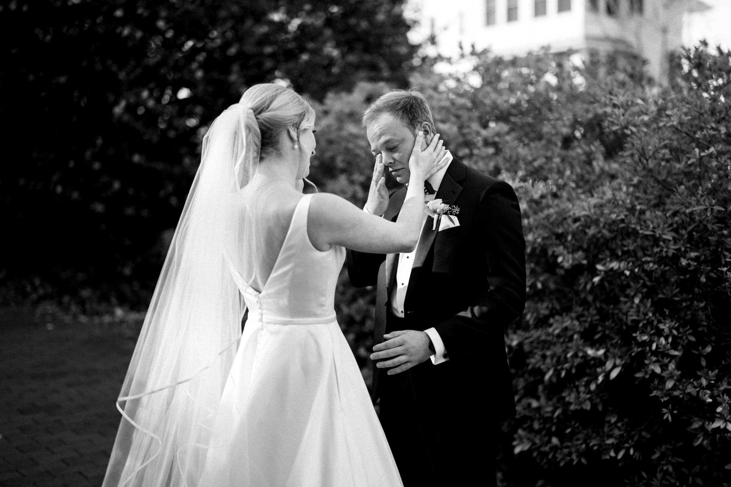 A black and white photo of a bride and groom in an emotional moment. The bride, in a long dress and veil, gently holds the groom’s face. They stand outside the charming Pinehurst Resort, surrounded by lush greenery, with an elegant building visible in the background.