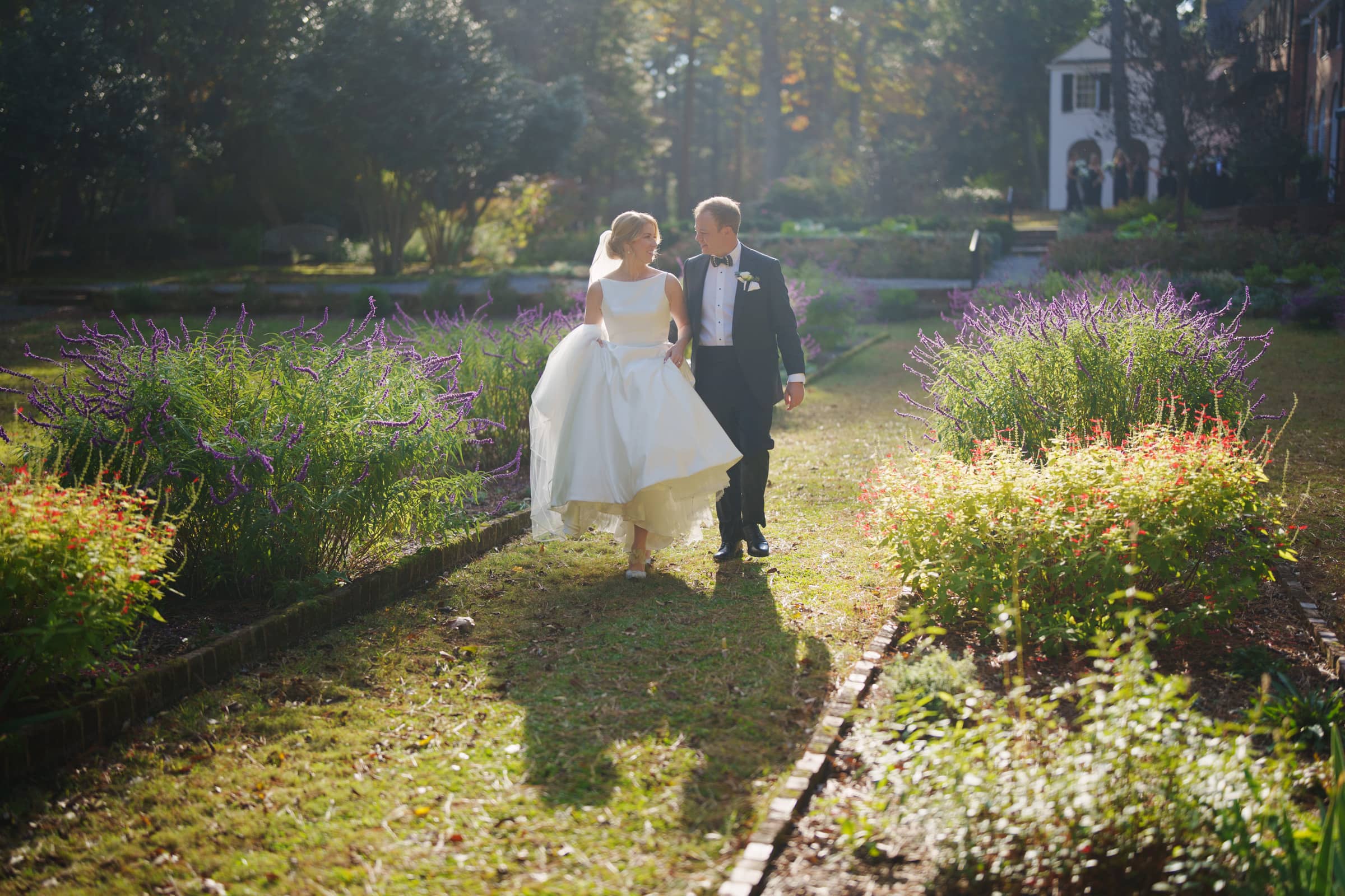 A bride and groom walk hand in hand through a sunlit garden filled with purple and red flowers at the charming Pinehurst Resort. The bride wears a white gown, and the groom is in a dark suit. A large house is visible in the background amidst tall trees.
