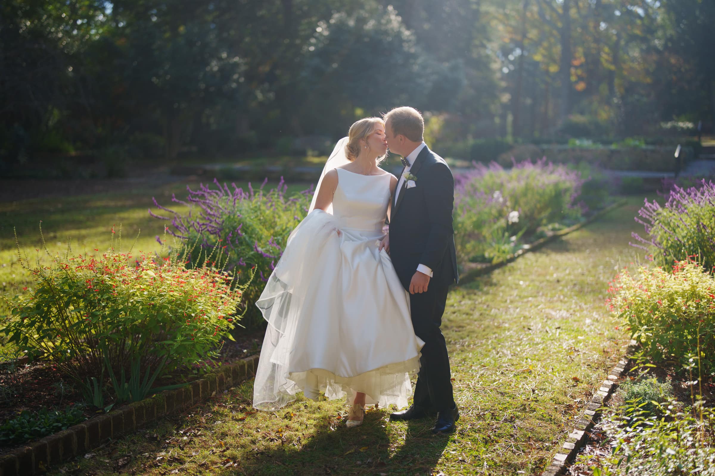 In a sunlit garden near the village chapel, a bride and groom share a kiss. The bride wears a flowing white dress, and the groom dons a dark suit. They're enveloped by lush foliage and purple flowers as sunlight filters through the trees, creating an enchanting scene reminiscent of Pinehurst Resort.