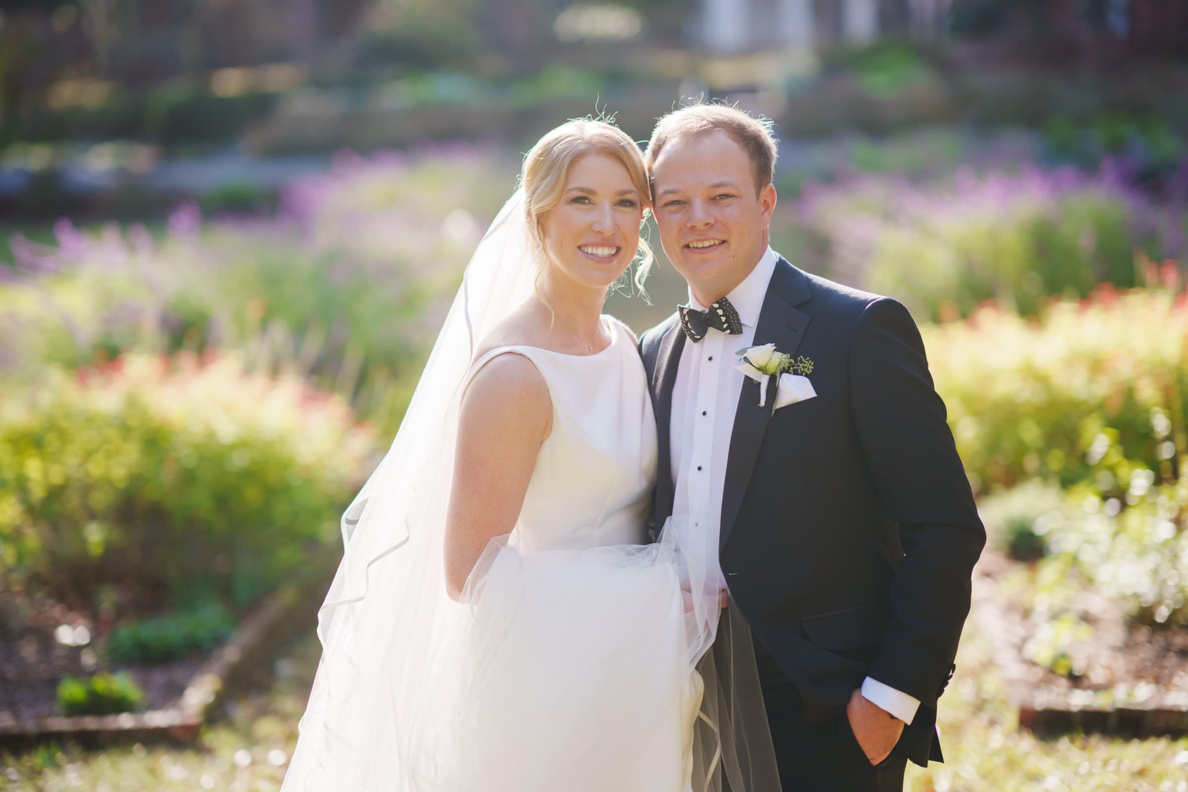 A bride in a white dress and veil stands next to a groom in a dark suit with a bow tie. They are smiling and posing outside at Pinehurst Resort, surrounded by vibrant flowers, capturing their perfect moment before heading to the charming village chapel for their ceremony.