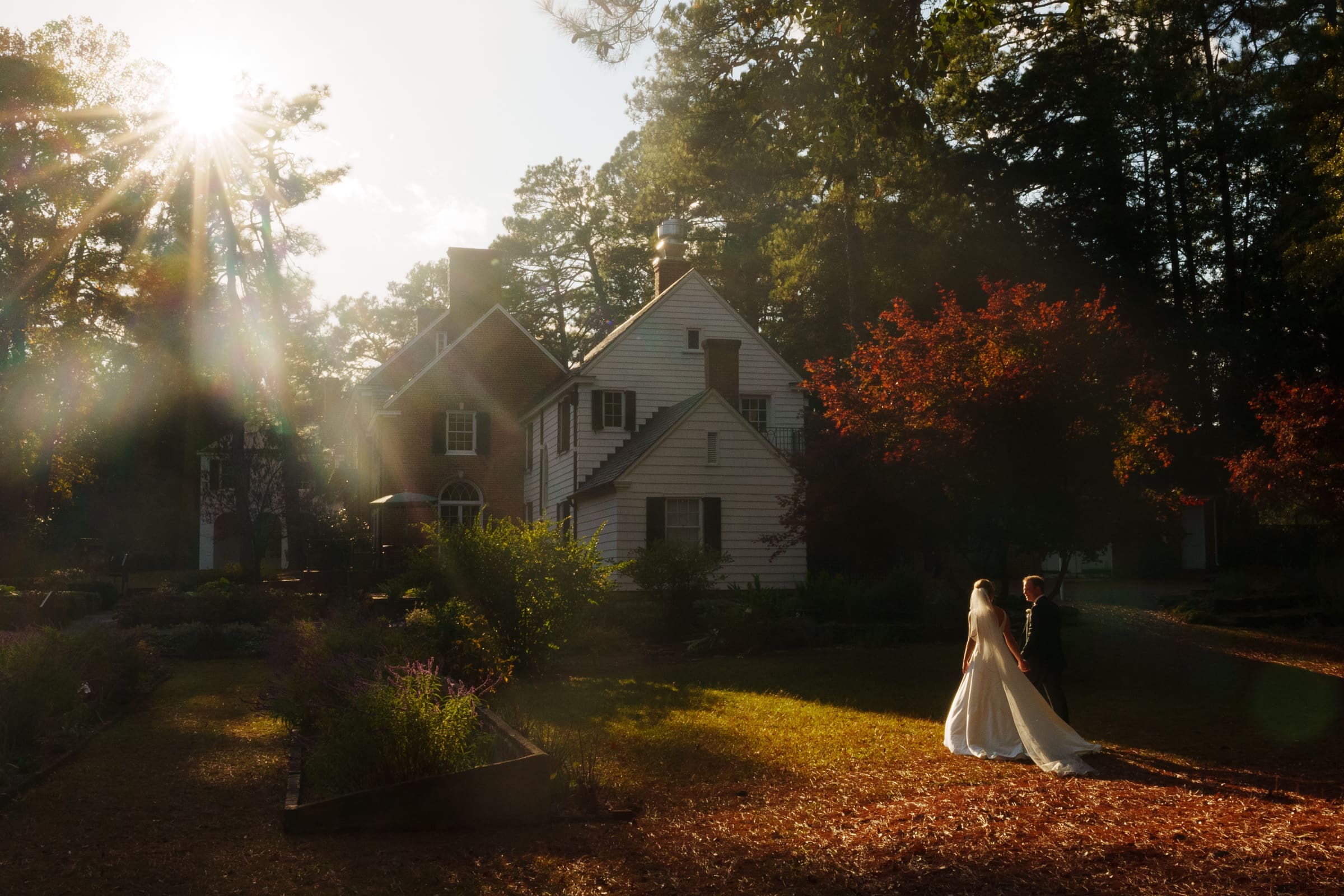 A couple in wedding attire stands before a charming house resembling a village chapel, nestled amidst lush greenery and tall trees. Sunlight filters through, casting a warm glow that echoes the enchantment of Pinehurst Resort.