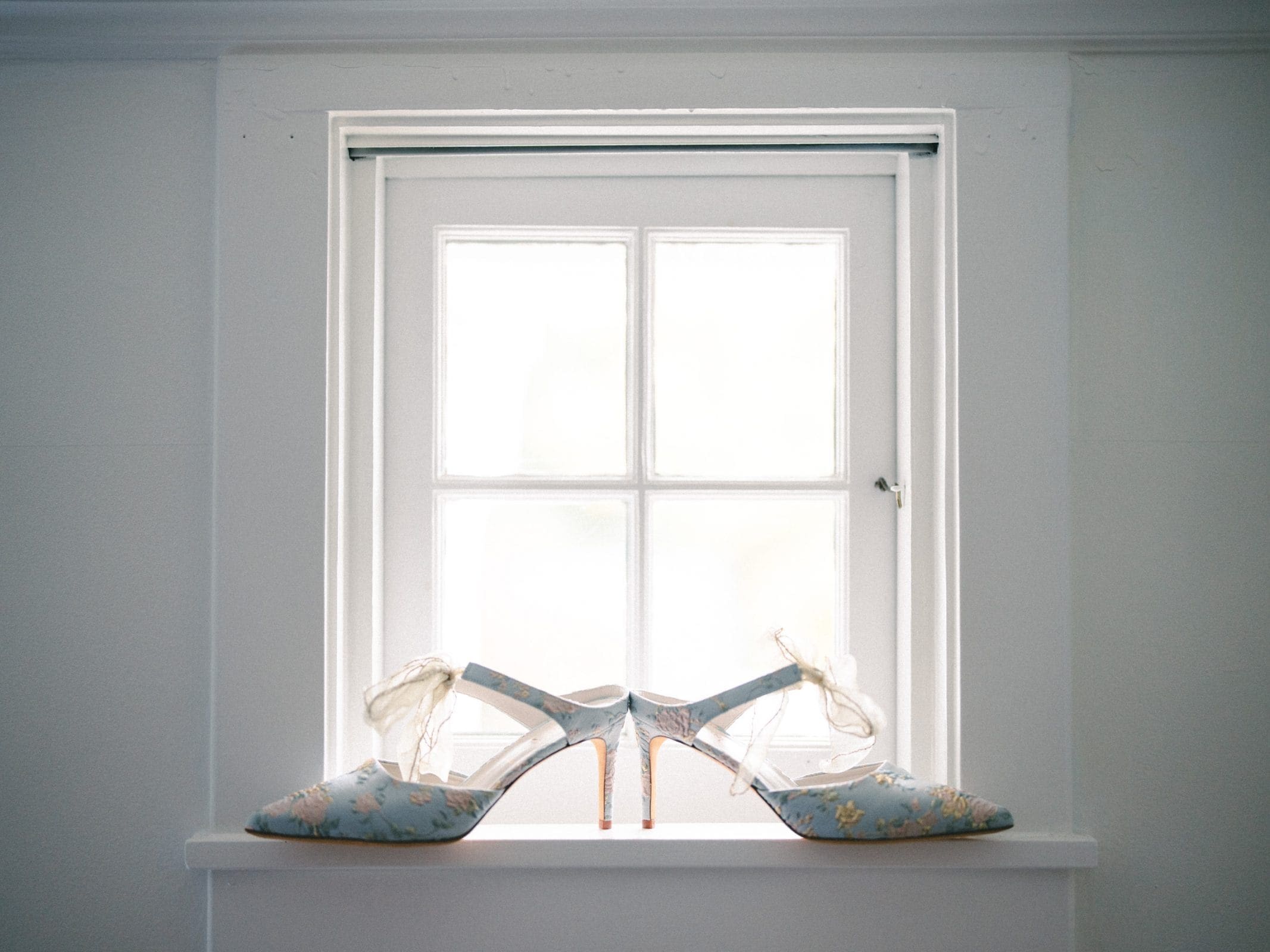 A pair of elegant, floral-patterned heels with delicate ankle ribbons is displayed on a windowsill at Pinehurst Resort, silhouetted against a softly lit window. The shoes have a light blue hue with intricate gold accents, creating a romantic and sophisticated ambiance.