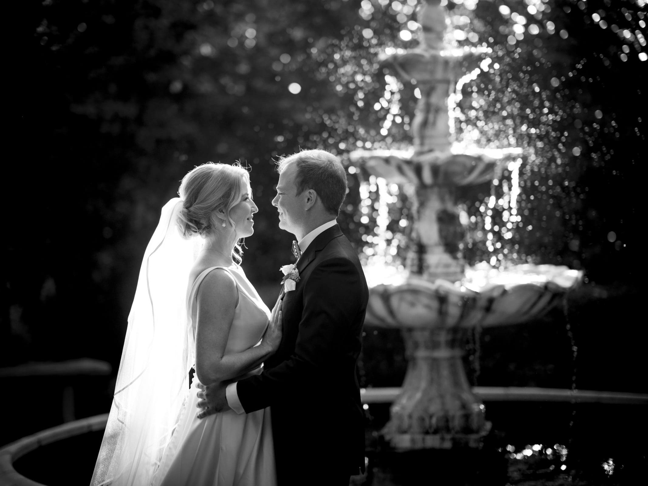 A bride and groom stand in front of a cascading fountain at Pinehurst Resort. The sunlight creates a glowing effect around them, highlighting the bride’s veil and their embrace. The scene is as romantic and intimate as a wedding in a quaint village chapel.