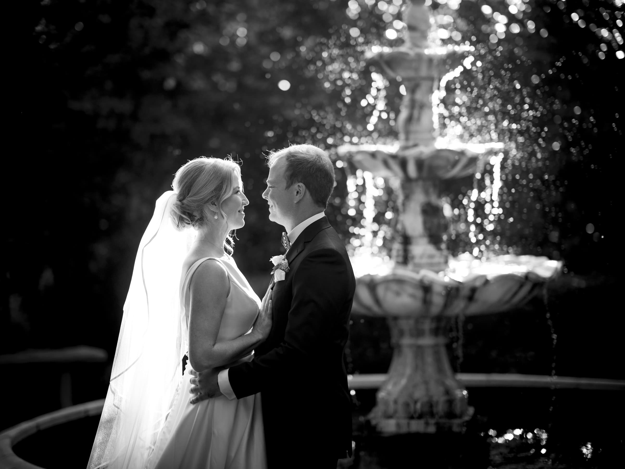 A bride and groom stand in front of a cascading fountain at Pinehurst Resort. The sunlight creates a glowing effect around them, highlighting the bride’s veil and their embrace. The scene is as romantic and intimate as a wedding in a quaint village chapel.