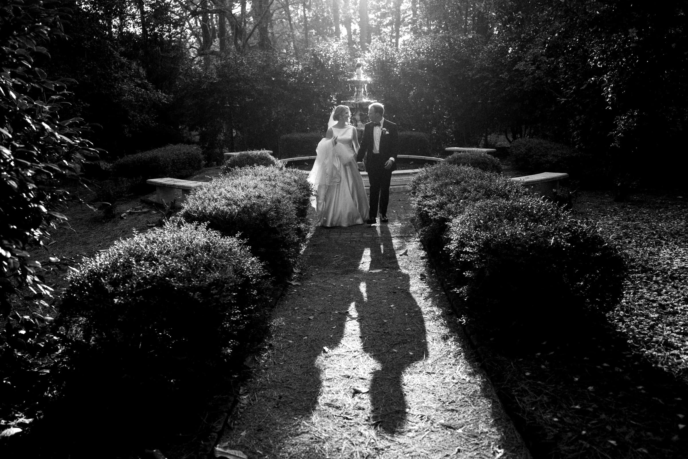 A bride and groom walk down a garden path in a sunlit wooded area, reminiscent of a tranquil village chapel, casting long shadows on the ground. Bushes line the path as sunlight filters through the trees at Pinehurst Resort, creating a serene and romantic scene.