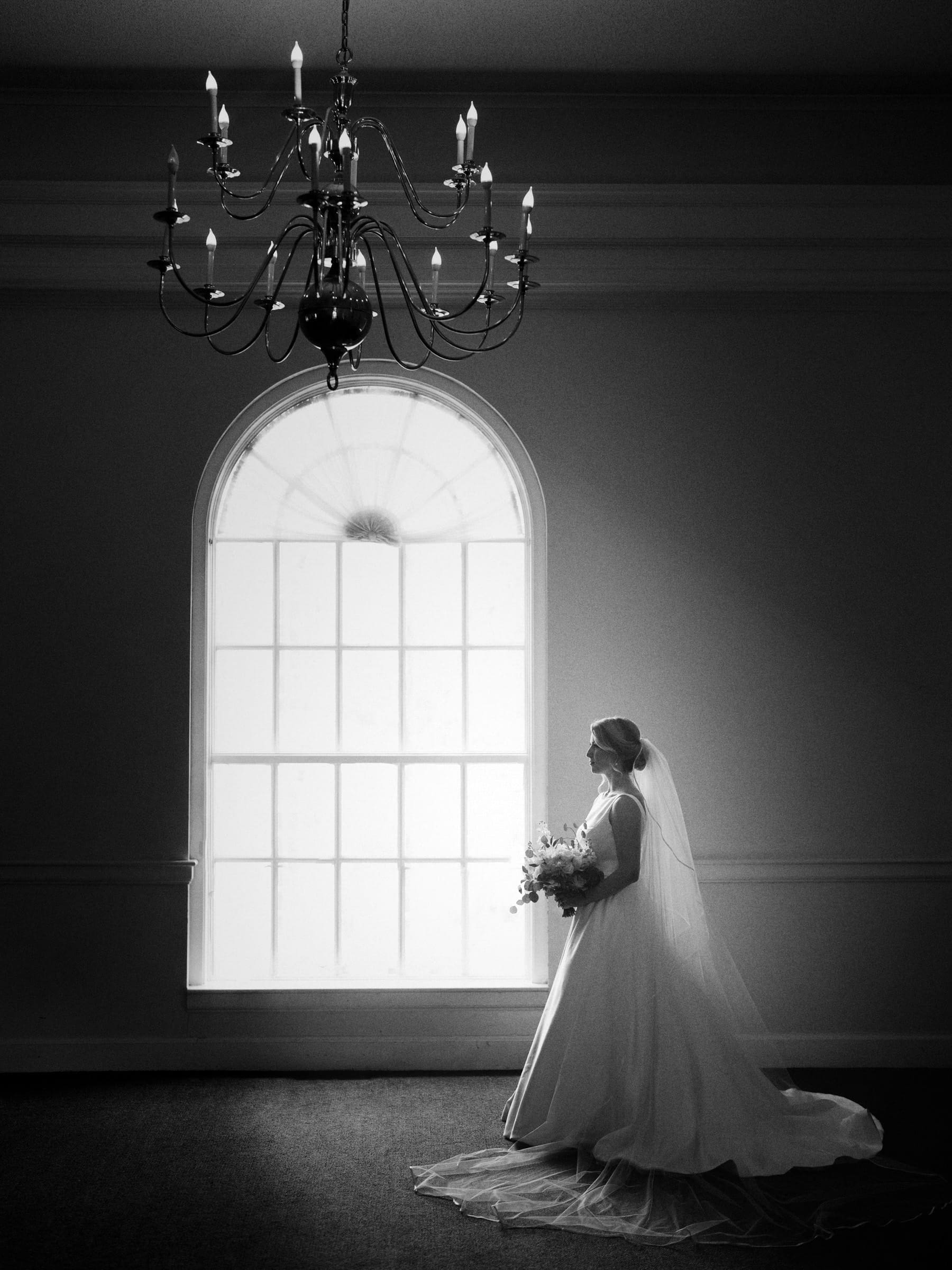 A bride in a flowing dress stands by a large arched window of the village chapel, holding a bouquet beneath the chandelier. The scene, captured in black and white at Pinehurst Resort, creates an elegant and timeless atmosphere.