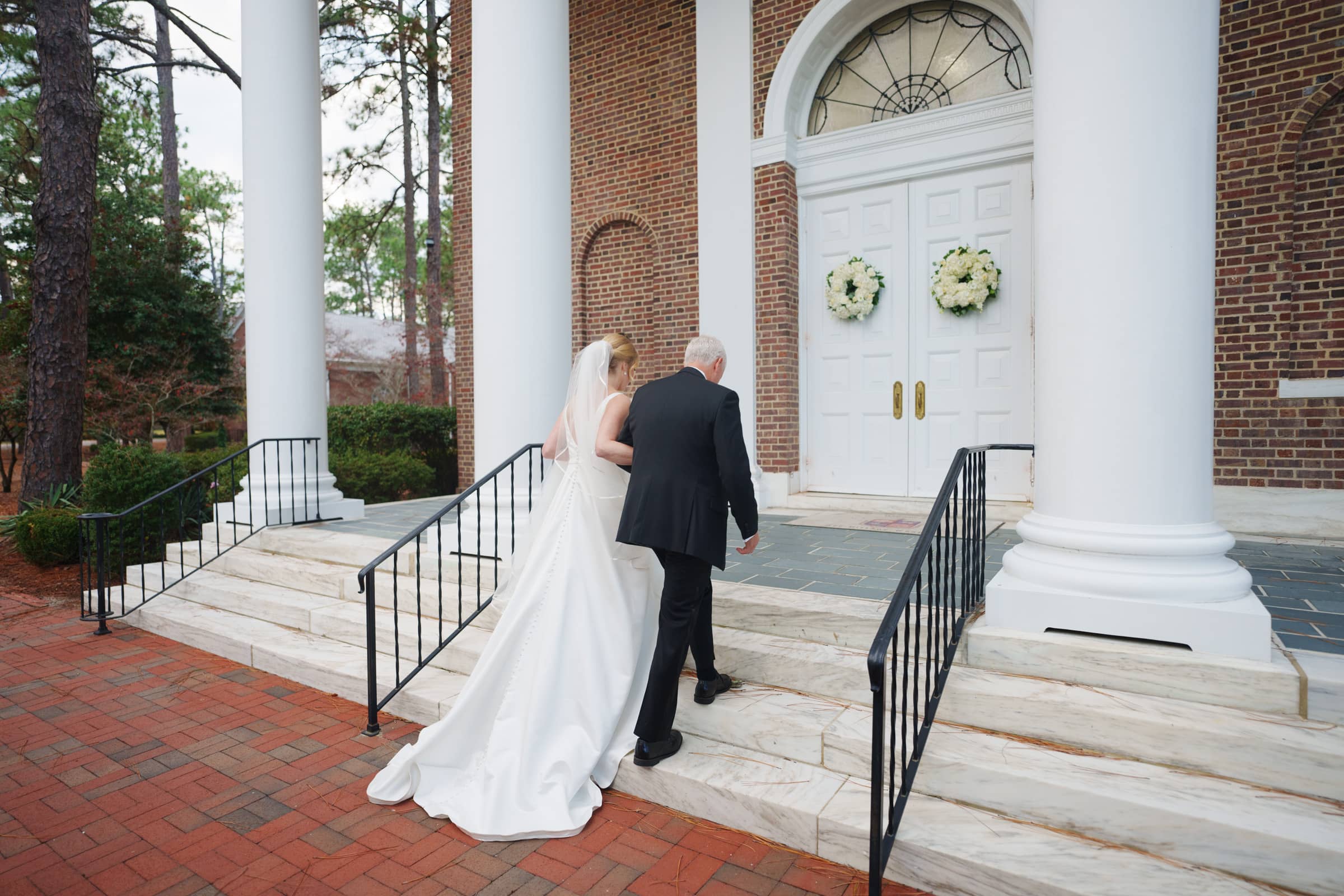 A bride in a white gown walks up the steps with an older man in a black suit, towards the charming village chapel with its brick facade, white columns, and double doors adorned with wreaths. This picturesque setting provides a perfect backdrop for their formal occasion.