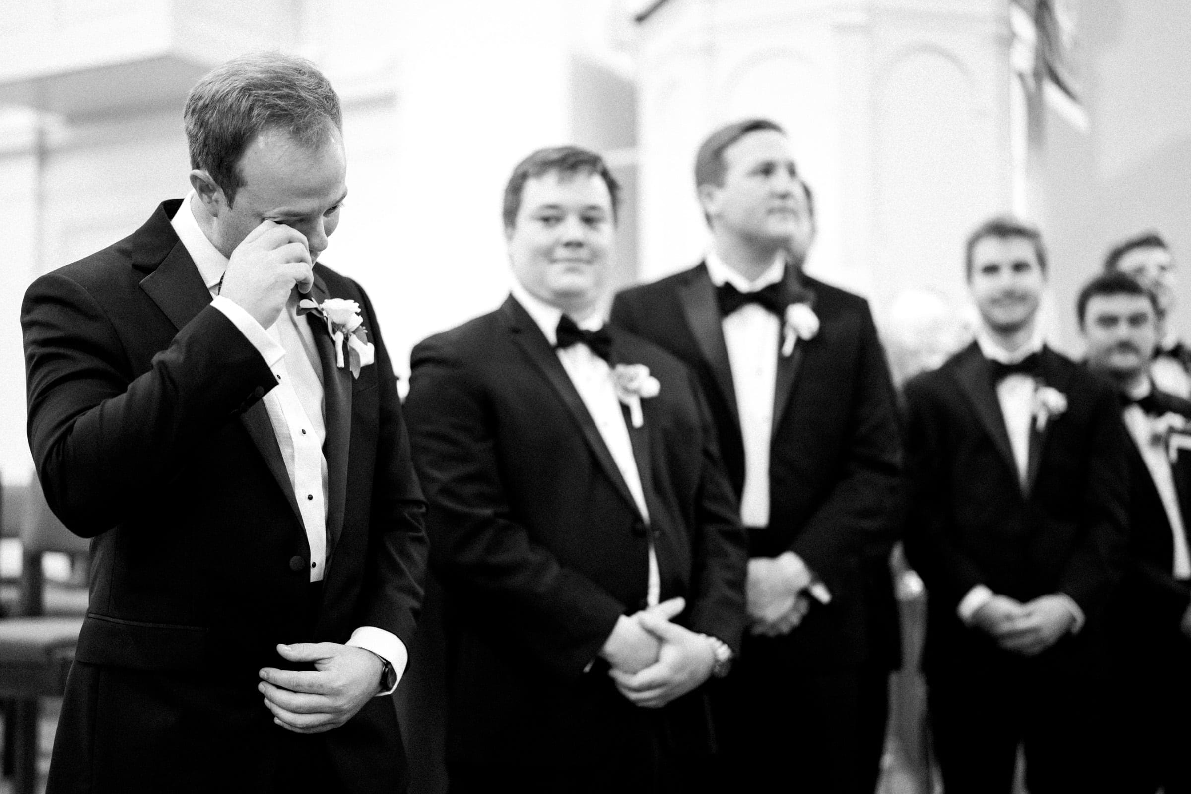A group of men dressed in black suits and bow ties stand together indoors, evoking the elegance of a Pinehurst Resort gathering. The man on the left appears emotional and wipes his eye, holding a paper. The background shows architectural details and a U.S. flag, enhancing the formal atmosphere.