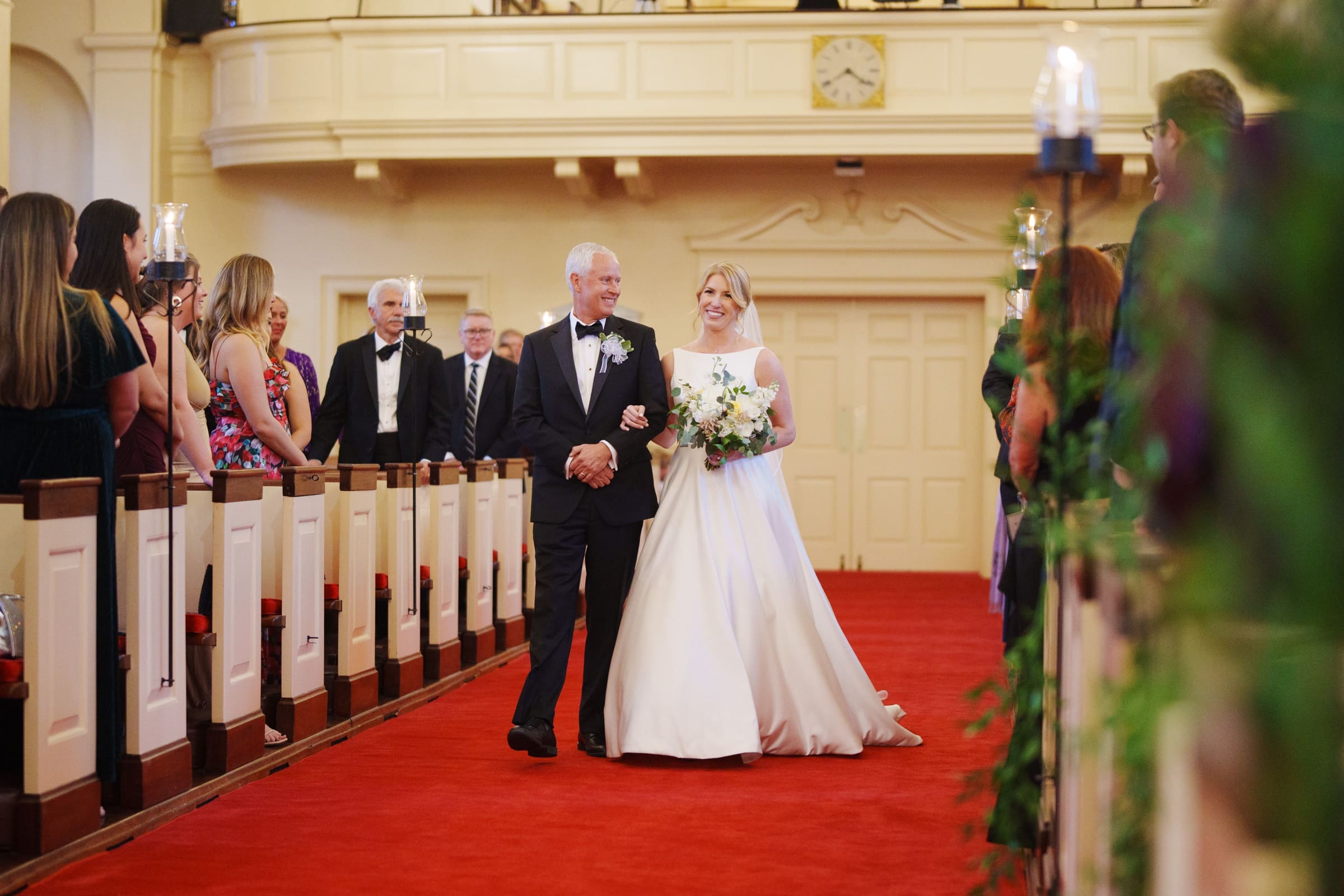 A bride in a white gown walks down the aisle of a charming village chapel with an older man in a black tuxedo. Guests line the red carpet, and a clock hangs on the wall above the entrance, creating an idyllic setting reminiscent of Pinehurst Resort's timeless elegance.