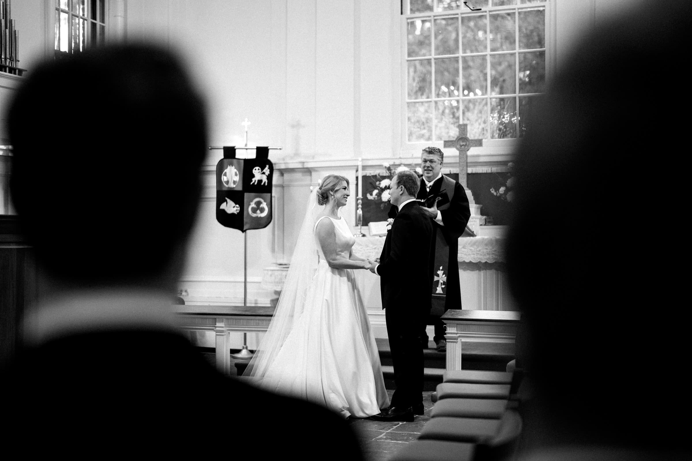 In a quaint village chapel, a bride and groom exchange vows with a pastor officiating. They stand at the altar of the serene Pinehurst Resort, holding hands amidst soft lighting and floral decorations. Silhouetted figures observe from the foreground, adding to the intimate ambiance.