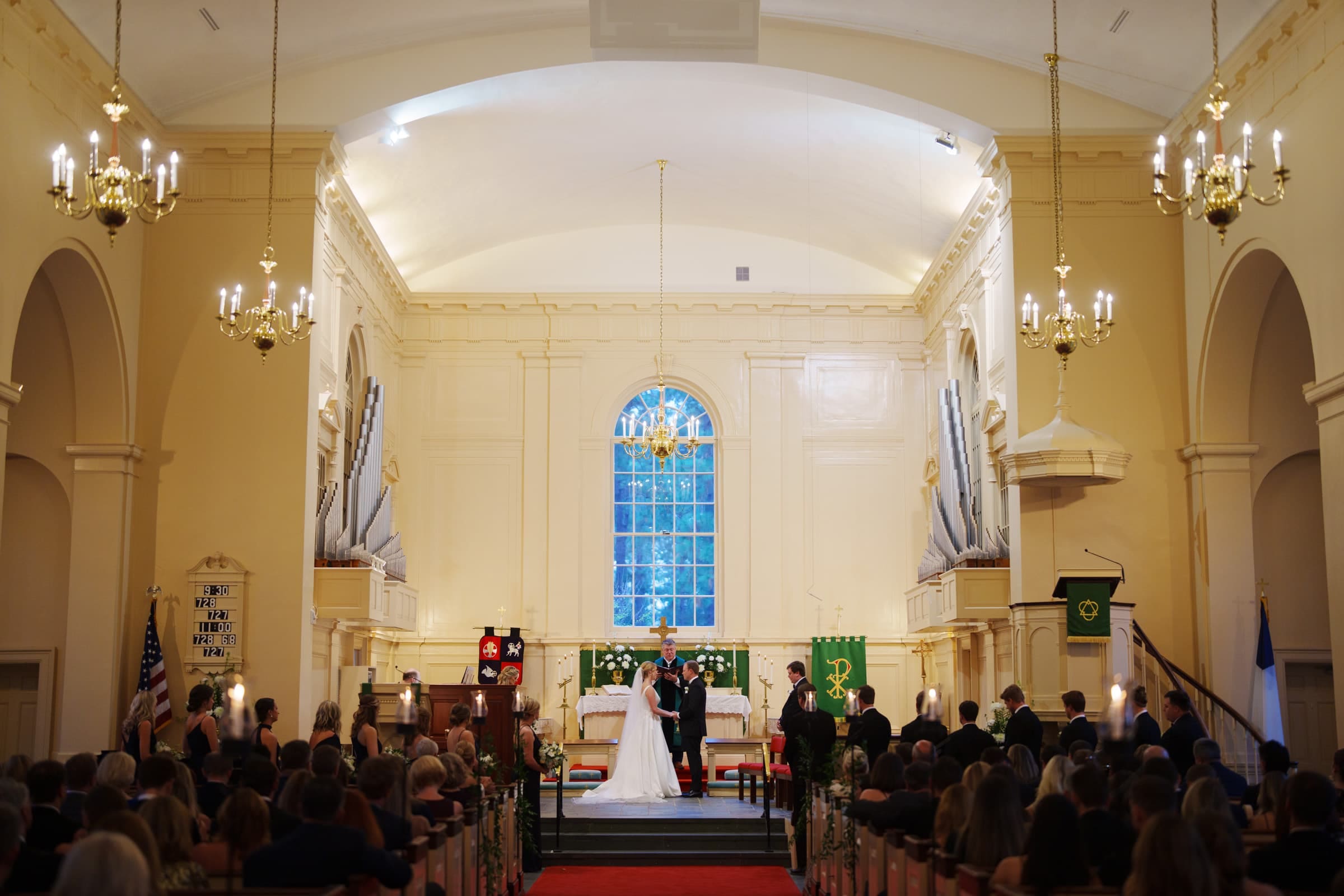 A couple stands at the altar in a charming village chapel for their wedding ceremony. Guests are seated in pews, and chandeliers hang from the high arched ceiling, creating a formal and elegant atmosphere reminiscent of Pinehurst Resort's classic charm.