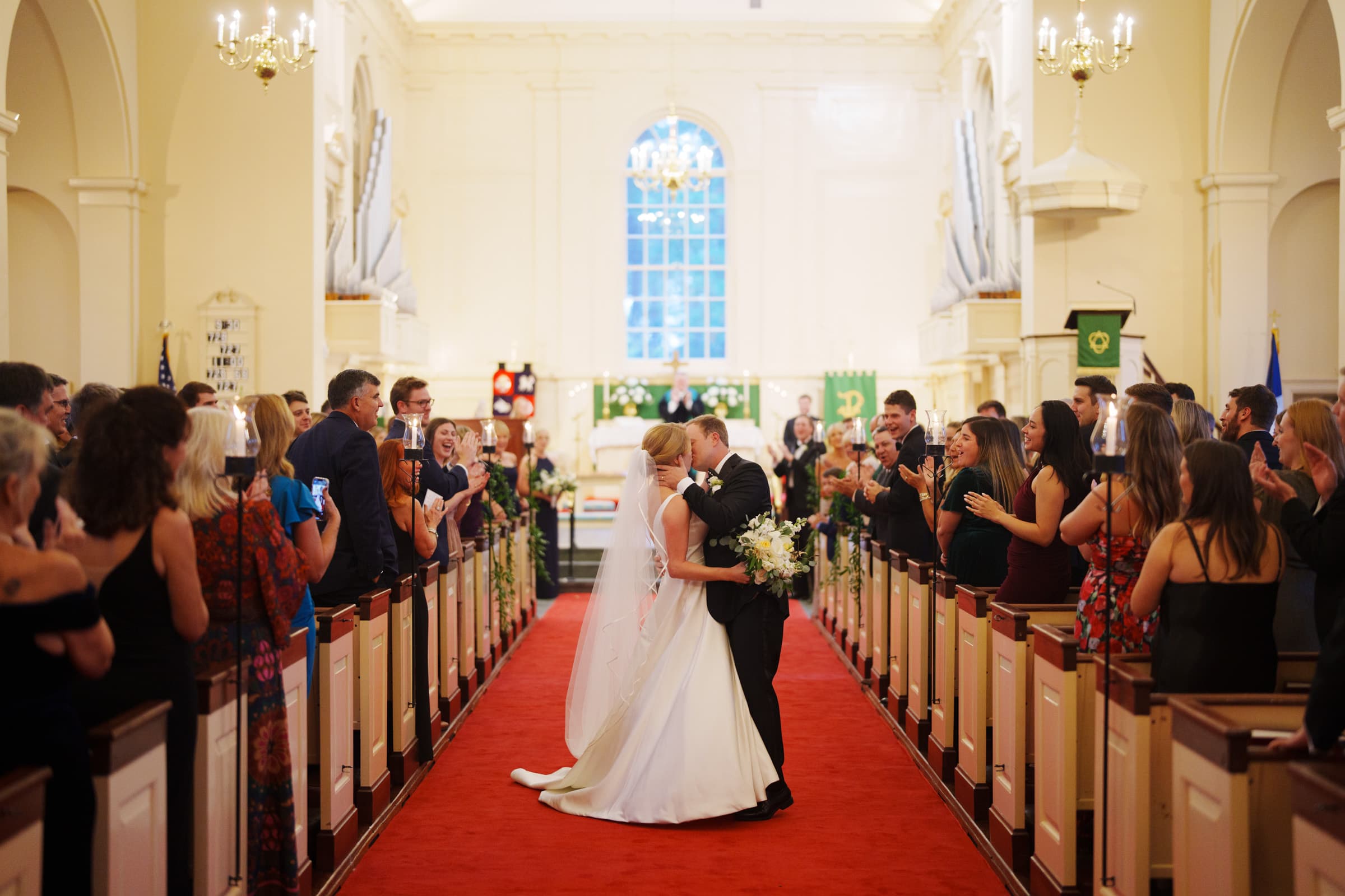 A newlywed couple kisses at the altar of a beautifully decorated village chapel. They are surrounded by a large group of applauding guests. The bride holds a bouquet, and the red carpet leads down the aisle with grand windows in the background.
