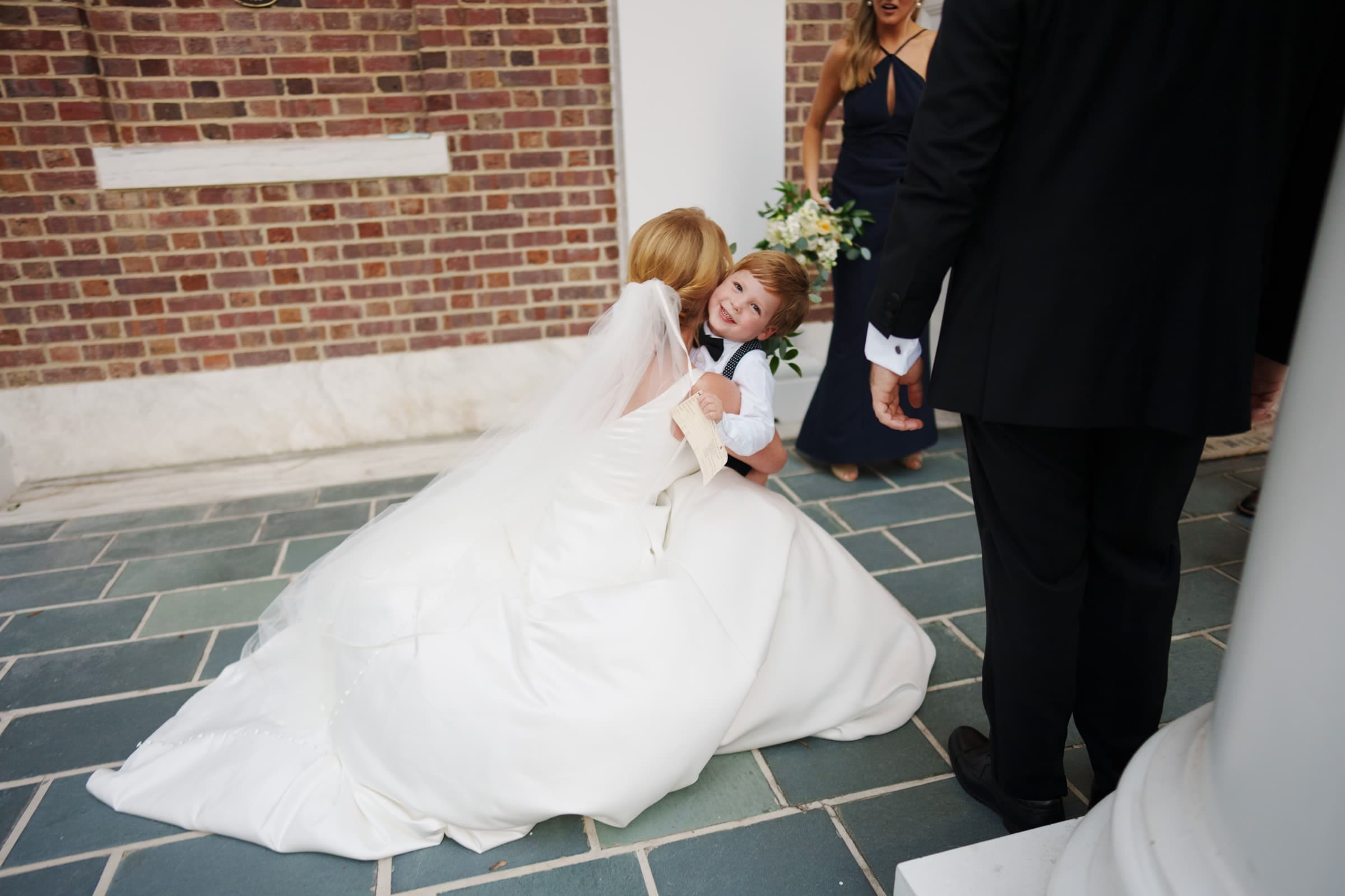 A bride in a white gown kneels to hug a young boy who is smiling. They are on a tiled outdoor area next to a brick wall reminiscent of the charming village chapel. Partially visible people in formal attire stand nearby, adding to the picturesque scene.