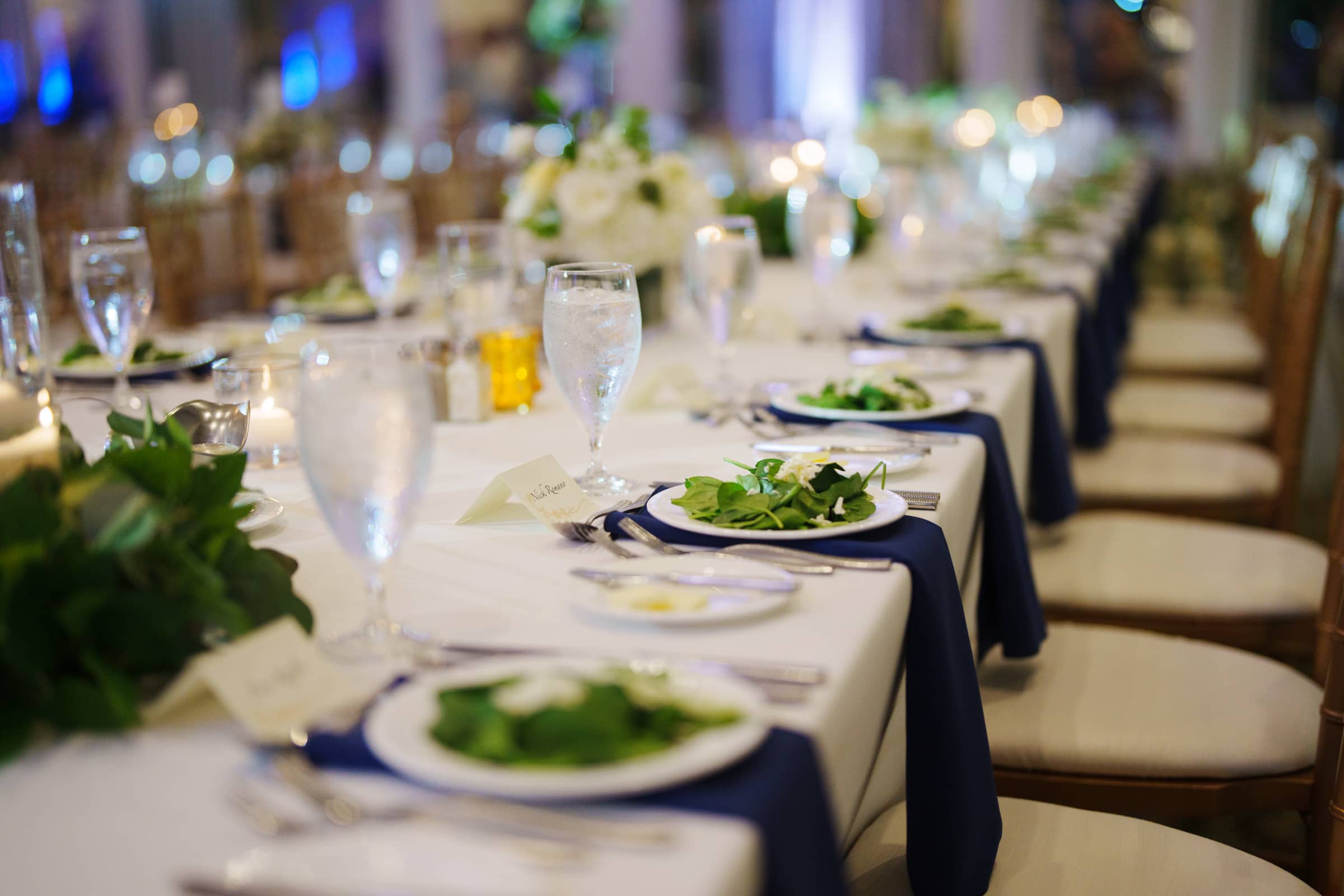 A long, elegantly set dining table at the village chapel features white tablecloths and navy blue napkins. Each place setting includes a salad, glassware, and cutlery. Soft lighting and floral centerpieces create a refined atmosphere akin to the sophistication of Pinehurst Resort.