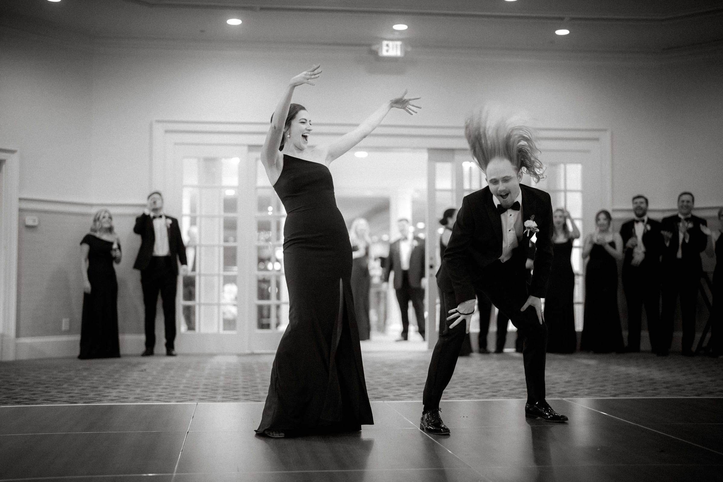 A black and white photo captures two people in formal attire dancing energetically on the village chapel's dance floor. The woman, in a long dress with arms raised, and the man, suited with long hair flying, are watched by onlookers in the background.