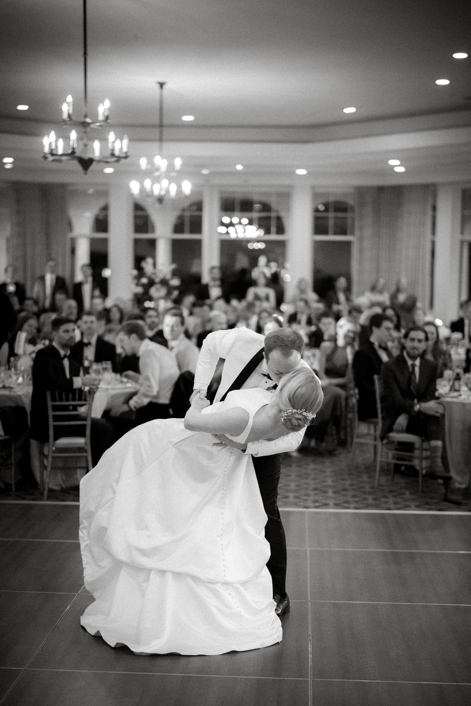 A couple shares a romantic dance on the elegant Pinehurst Resort dance floor, captivating seated guests. The venue, adorned with chandeliers and large windows, echoes the charm of a village chapel, creating a sophisticated atmosphere.
