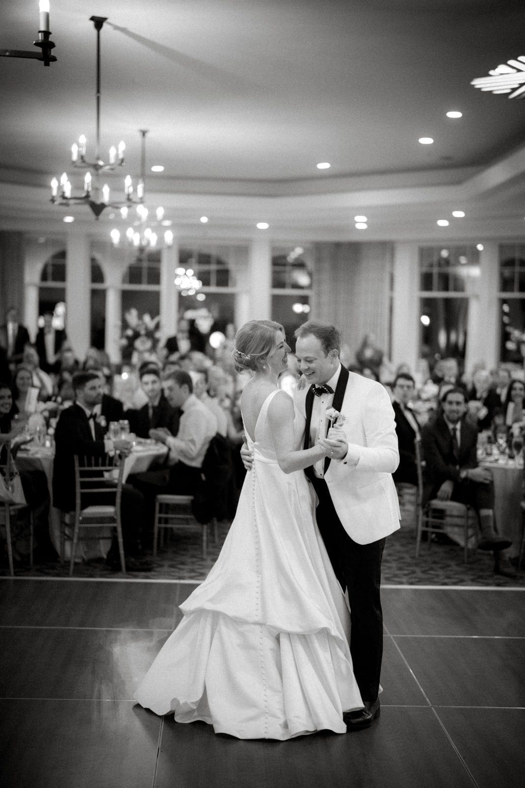 In a warmly lit room at Pinehurst Resort, a bride and groom share their first dance. The bride wears a flowing white gown, and the groom dons a white tuxedo jacket. Chandeliers illuminate the elegant setting filled with seated guests.