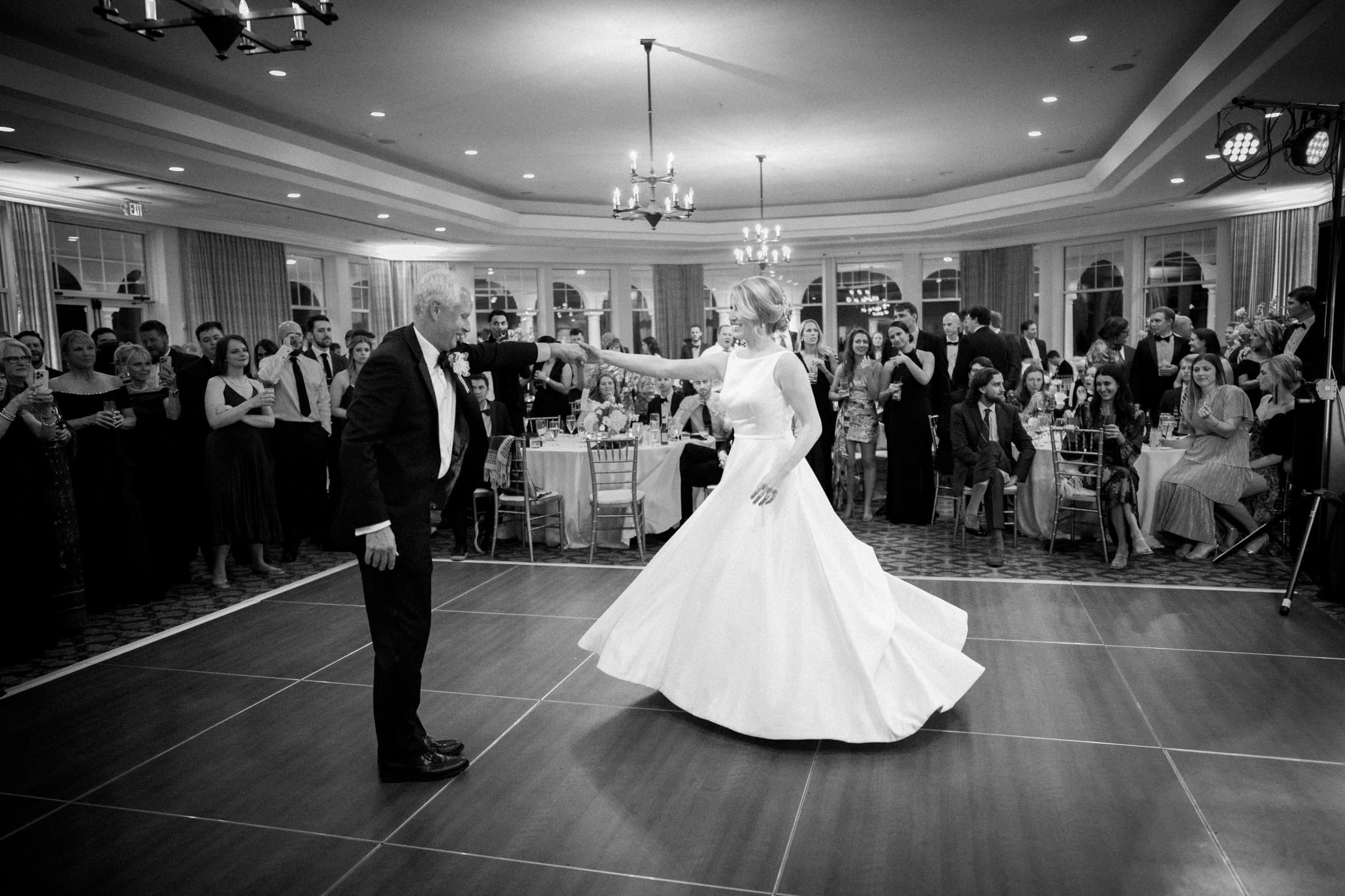 A bride and an older man, both in formal attire, dance on a wooden floor at the Pinehurst Resort. Guests seated and standing admire from afar as chandeliers and soft lighting create a warm atmosphere, reminiscent of a cozy village chapel.