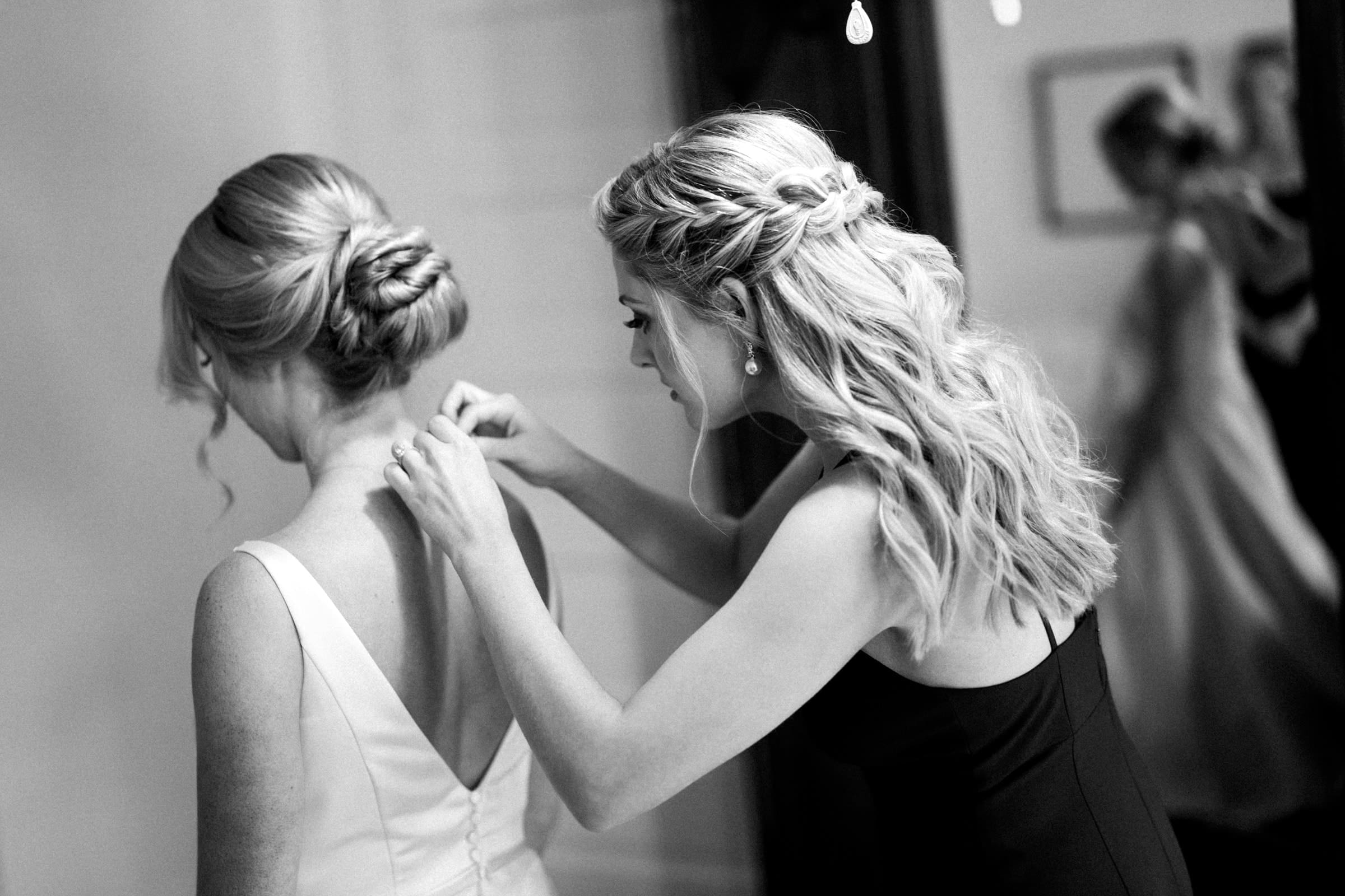 In a black and white photo, a woman assists another with her dress at the village chapel. The woman being helped has her back to the camera, both sporting styled hair. A softly blurred background, with a mirror reflecting part of this serene scene, adds depth to the moment.