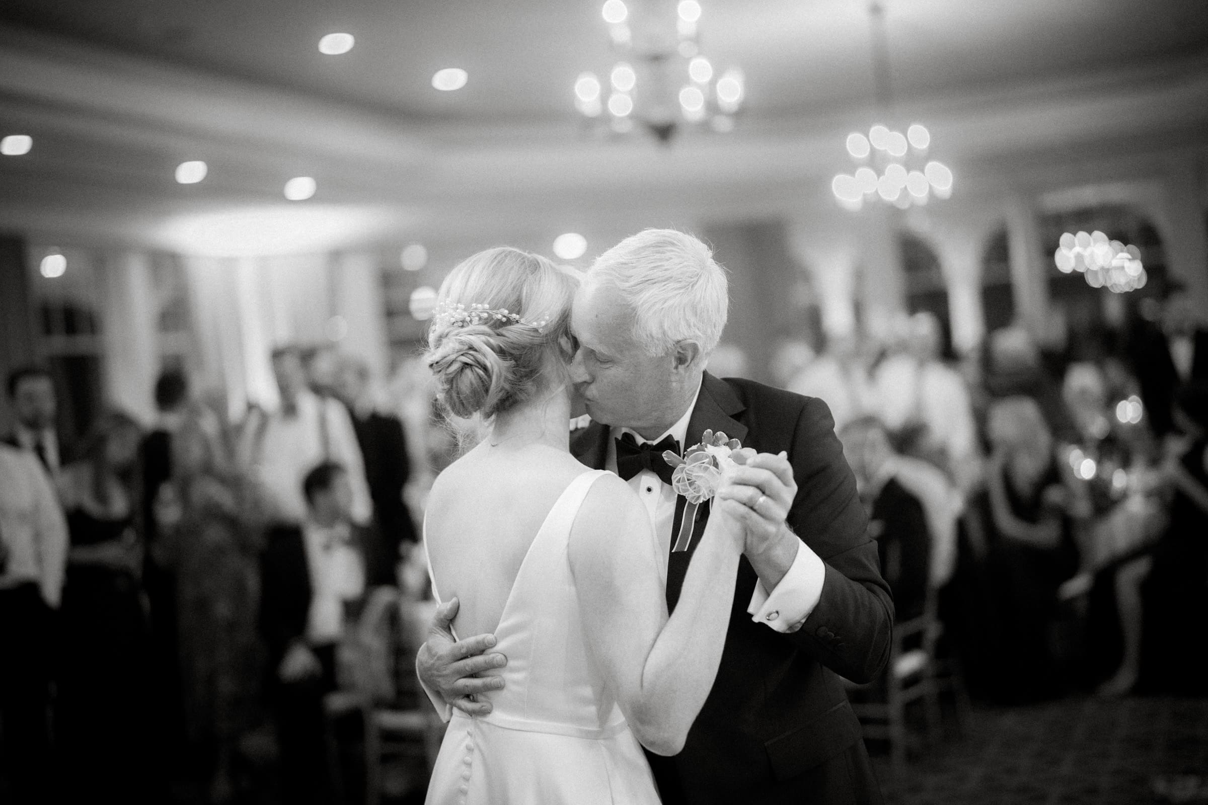 A man and a woman dance closely in a formal setting at the charming Pinehurst Resort, surrounded by elegantly dressed guests. The room is softly lit, and the atmosphere is joyful and celebratory.