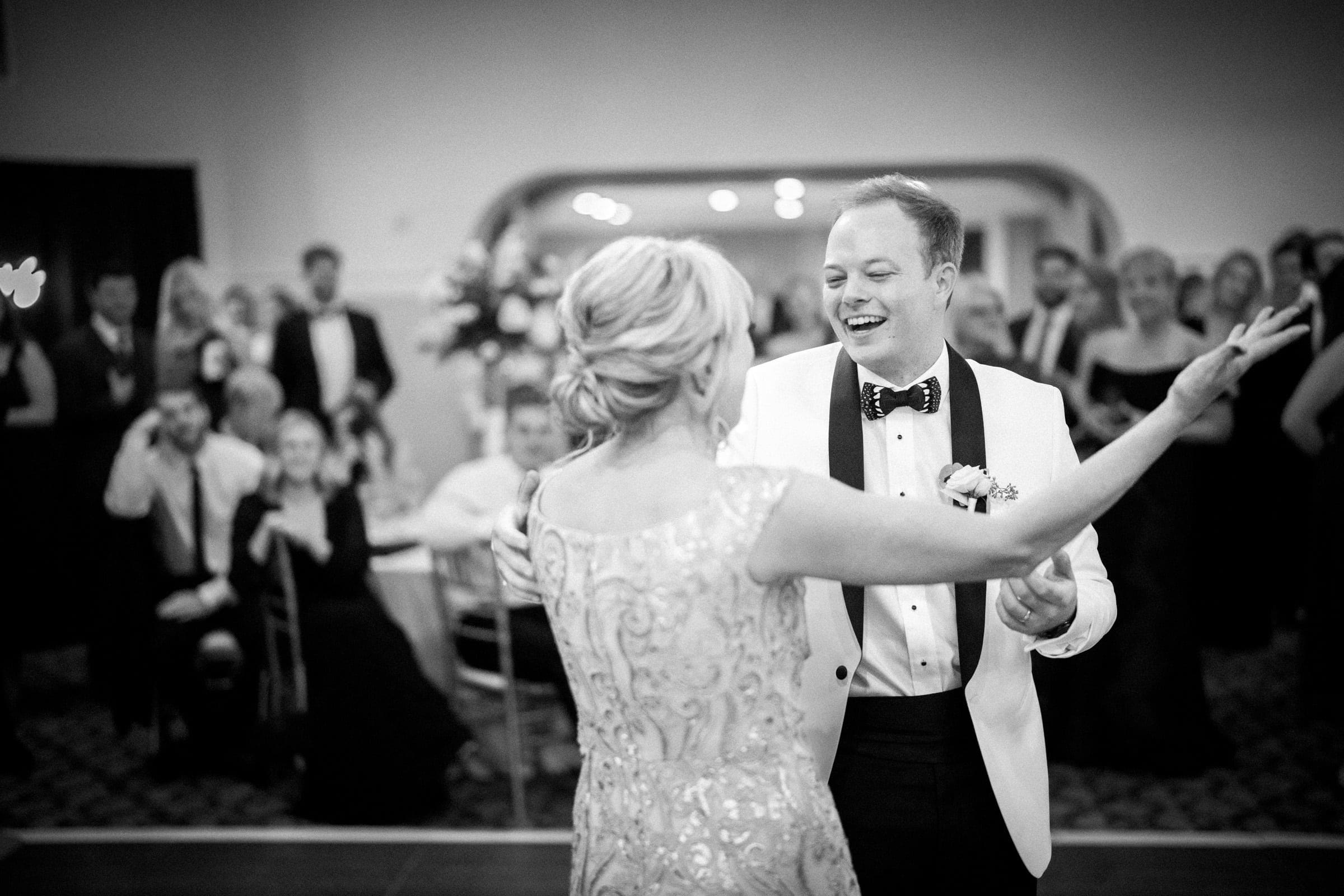 A black-and-white photo captures a couple dancing at a formal event, perhaps at the Pinehurst Resort. The man in a white suit and black bow tie smiles at the woman in an elegant dress. Guests in the background watch and smile.