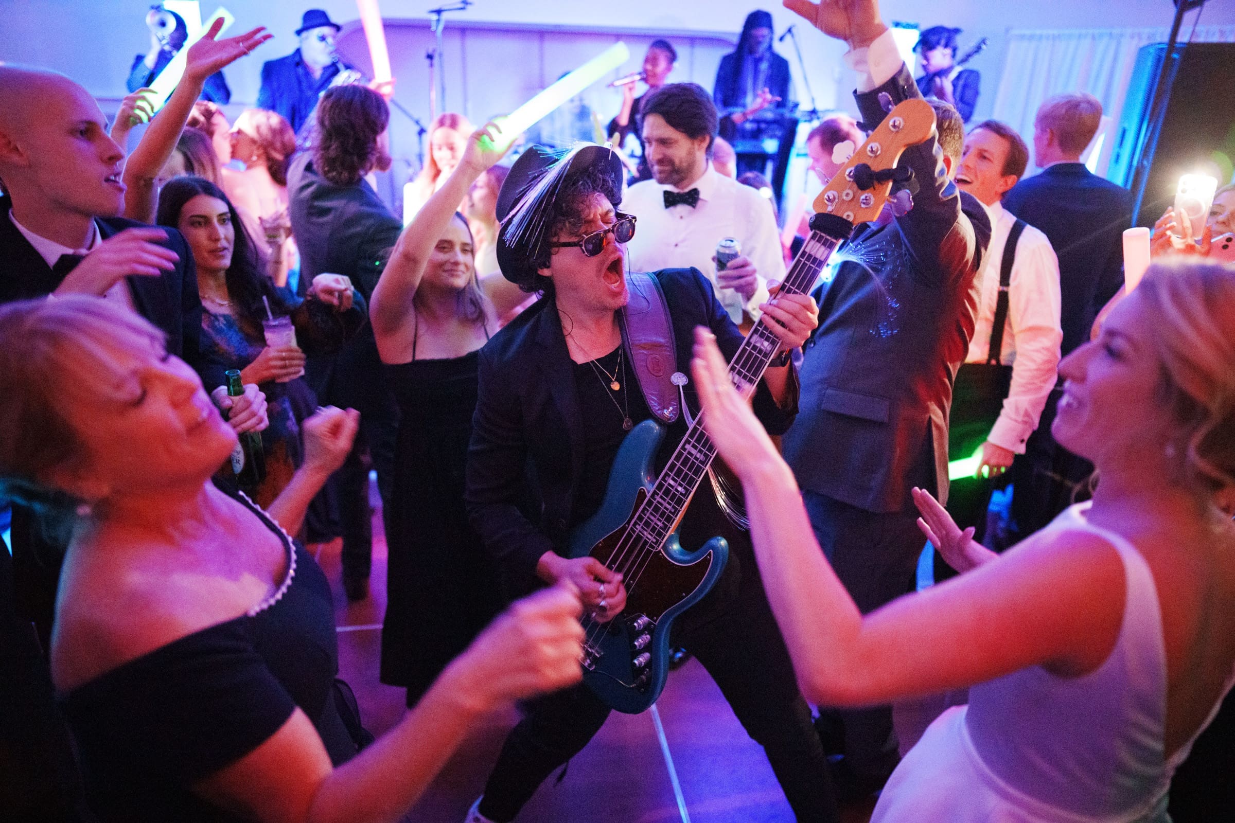In a lively party scene reminiscent of Pinehurst Resort, a musician energetically plays a bass guitar at the center, surrounded by people dancing and raising their hands. The atmosphere is vibrant, with colorful lights and a stage in the background.