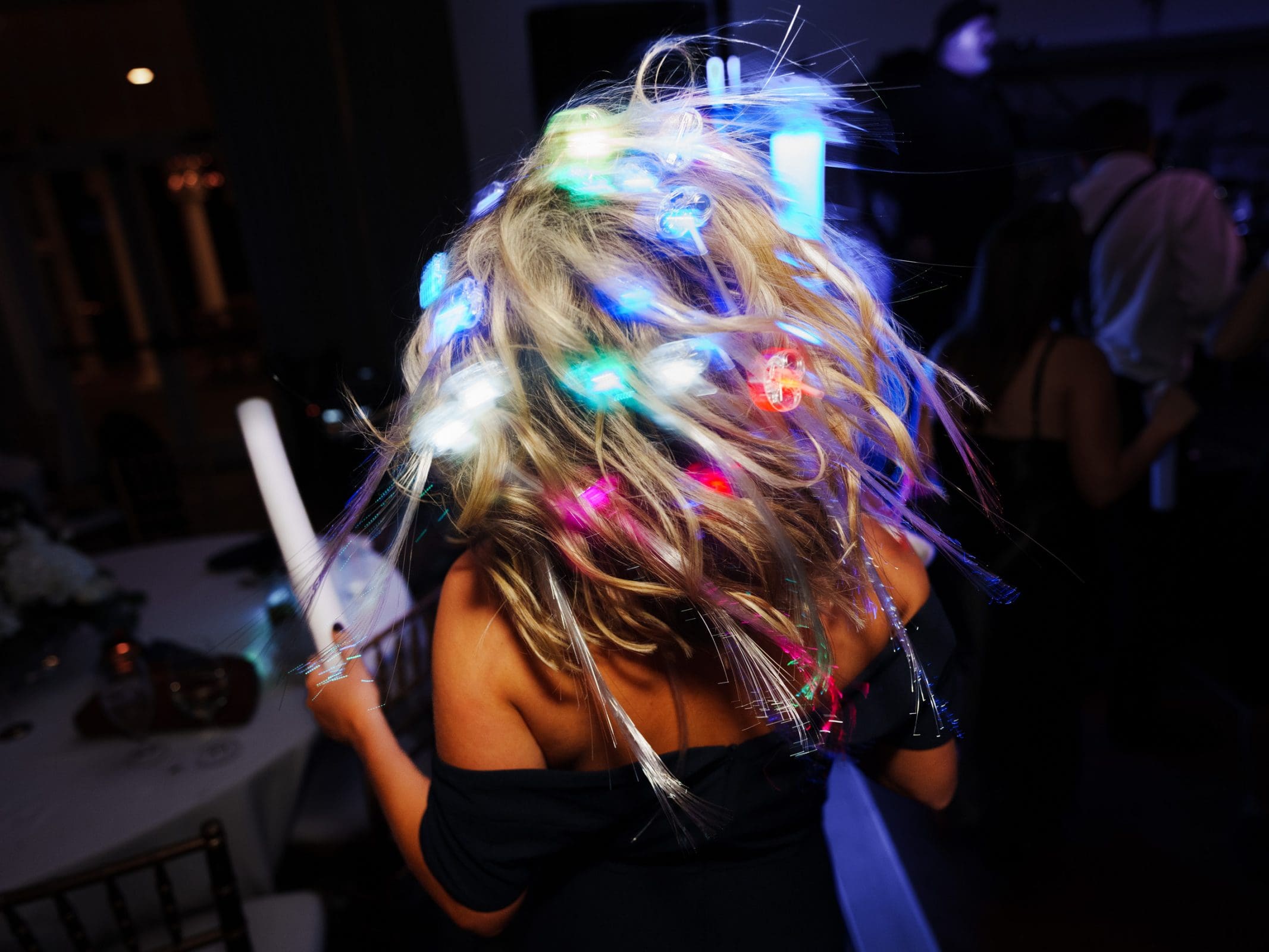 In a dark room, a person with blonde hair decorated with colorful LED lights twists and dances, creating a vibrant display against the dim setting. Reminiscent of a lively village chapel gathering, tables and chairs dot the background like those at Pinehurst Resort.