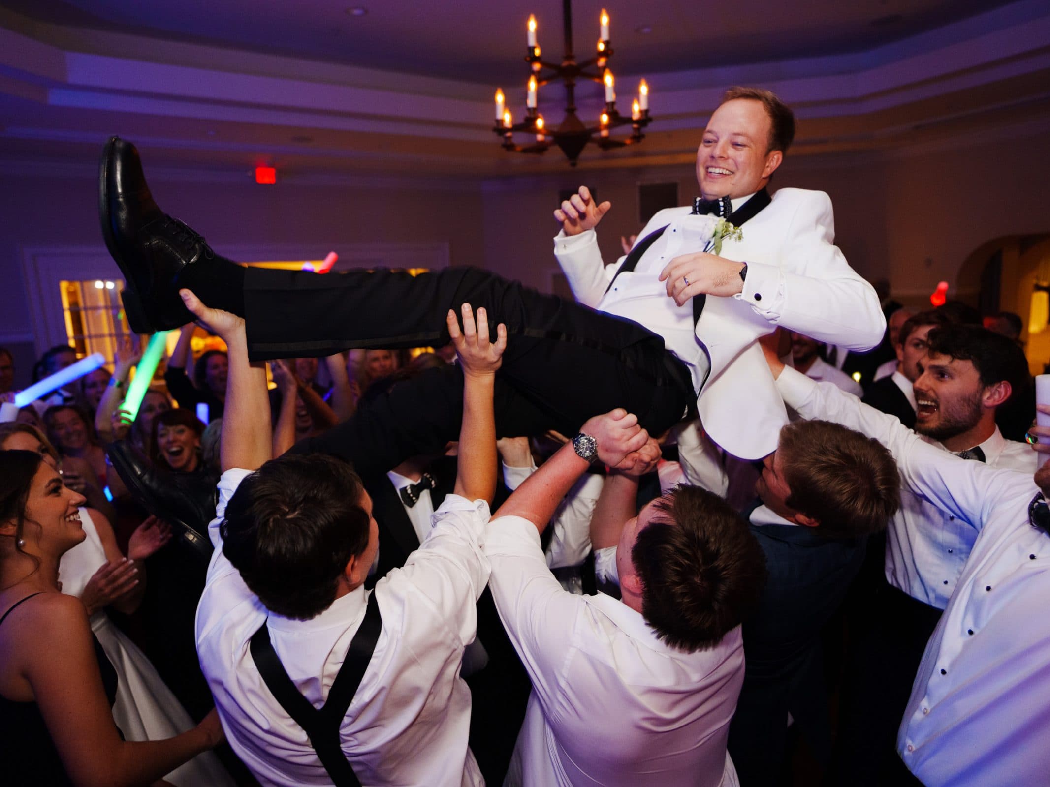 At a joyful wedding reception near the charming village chapel, the groom in a white tuxedo is lifted into the air by cheering guests. Some wave colorful glow sticks, filling the room with vibrant light as a chandelier sparkles above, capturing the enchanting spirit of Pinehurst Resort.