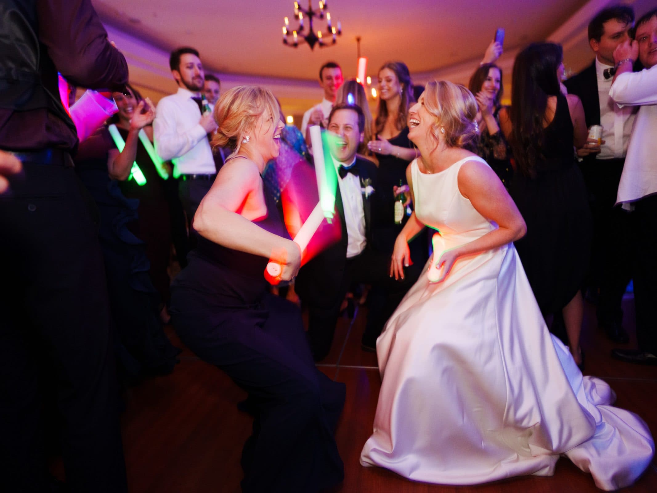 In a room elegantly decorated with chandeliers, a bride in a white dress and another woman dance energetically with colorful light sticks at the Pinehurst Resort. Surrounded by smiling guests, some holding phones, the scene is full of joy and celebration.