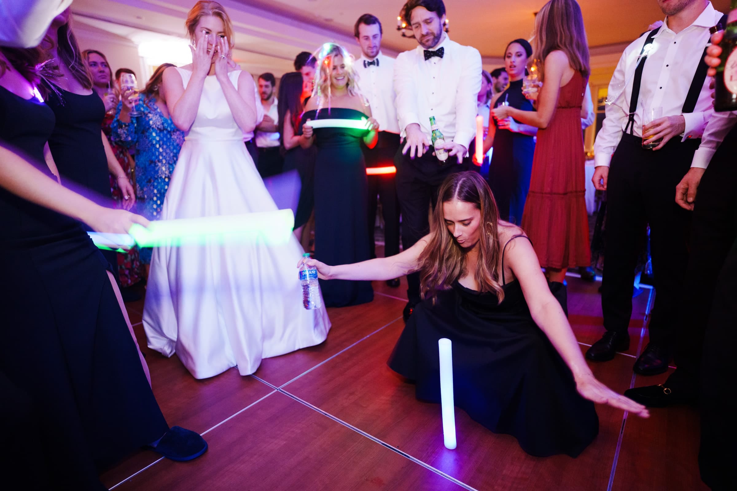 At a lively party scene, where a woman in a black dress energetically dances with glowing sticks on the floor, the atmosphere feels as vibrant as Pinehurst Resort. People in formal attire, including a bride in a white gown, watch with joy and surprise, reminiscent of celebrations at a quaint village chapel.