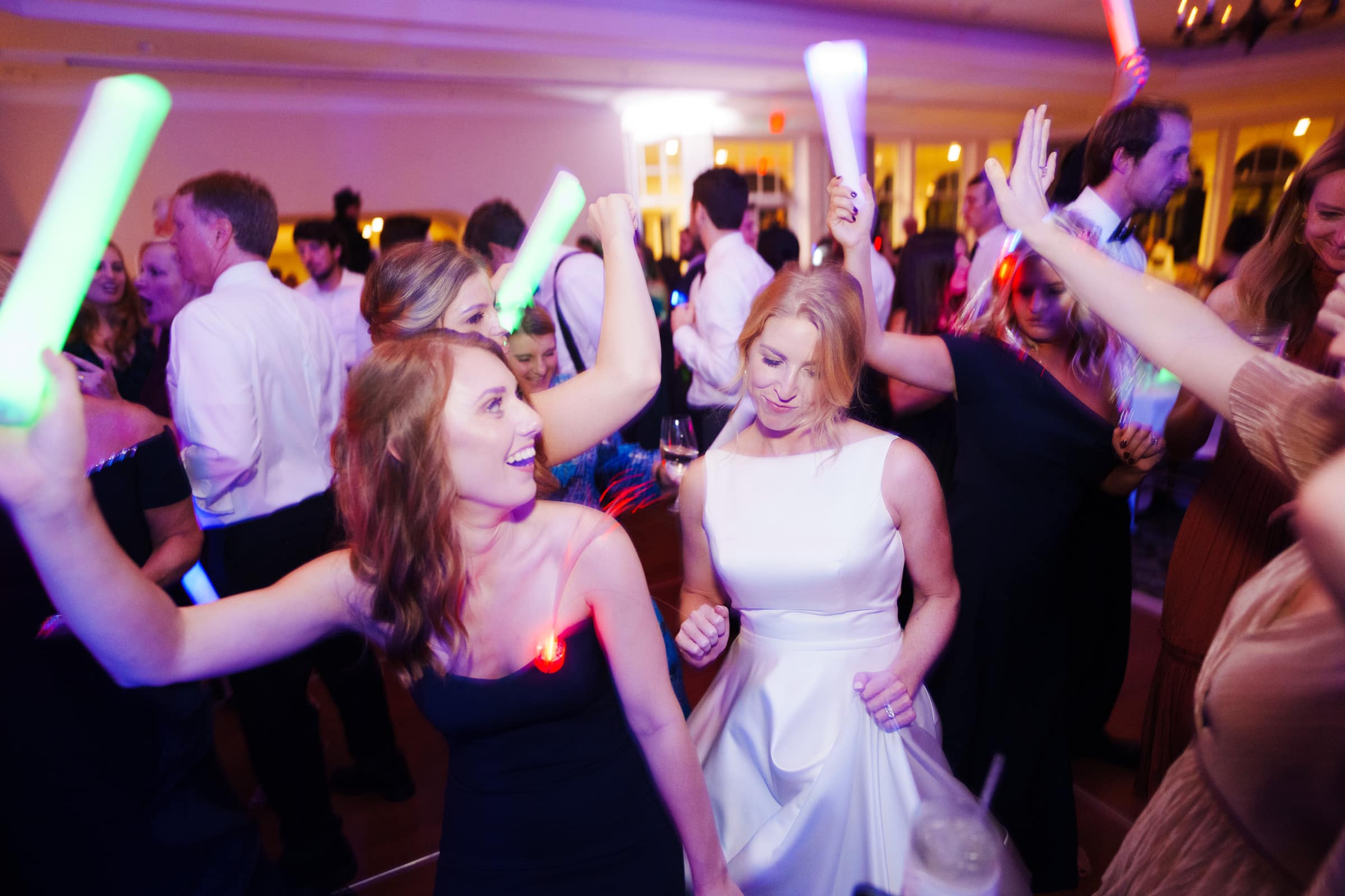 At a lively party in Pinehurst Resort, a group of people dances energetically, some holding colorful glowing sticks. Bright lighting illuminates their smiles as they move gracefully in formal attire, radiating joy and elegance.
