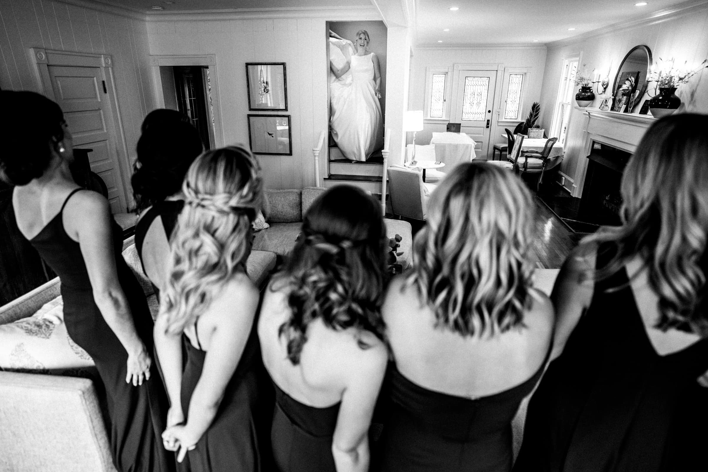 A bride stands on a staircase in a white dress, gazing at her bridesmaids lined up in black dresses in the elegantly decorated living room of Pinehurst Resort. The room features a cozy sofa, a fireplace, and framed pictures reminiscent of moments from their time at the village chapel.
