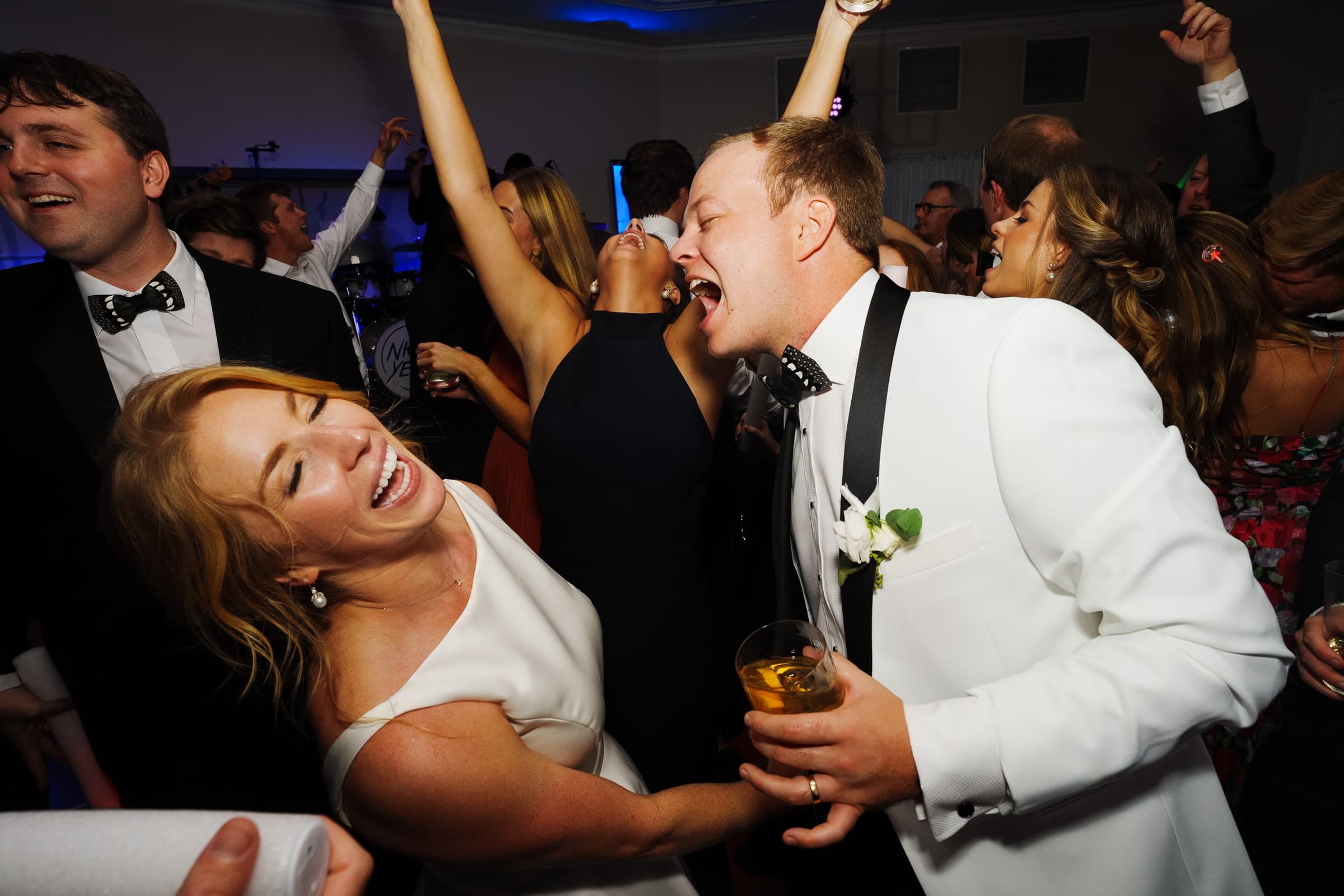 At the Pinehurst Resort, a lively celebration unfolds with people dancing and laughing. A man in a white tuxedo sings into a microphone, drink in hand. A woman in a white dress joyfully leans towards him, surrounded by cheerful guests.
