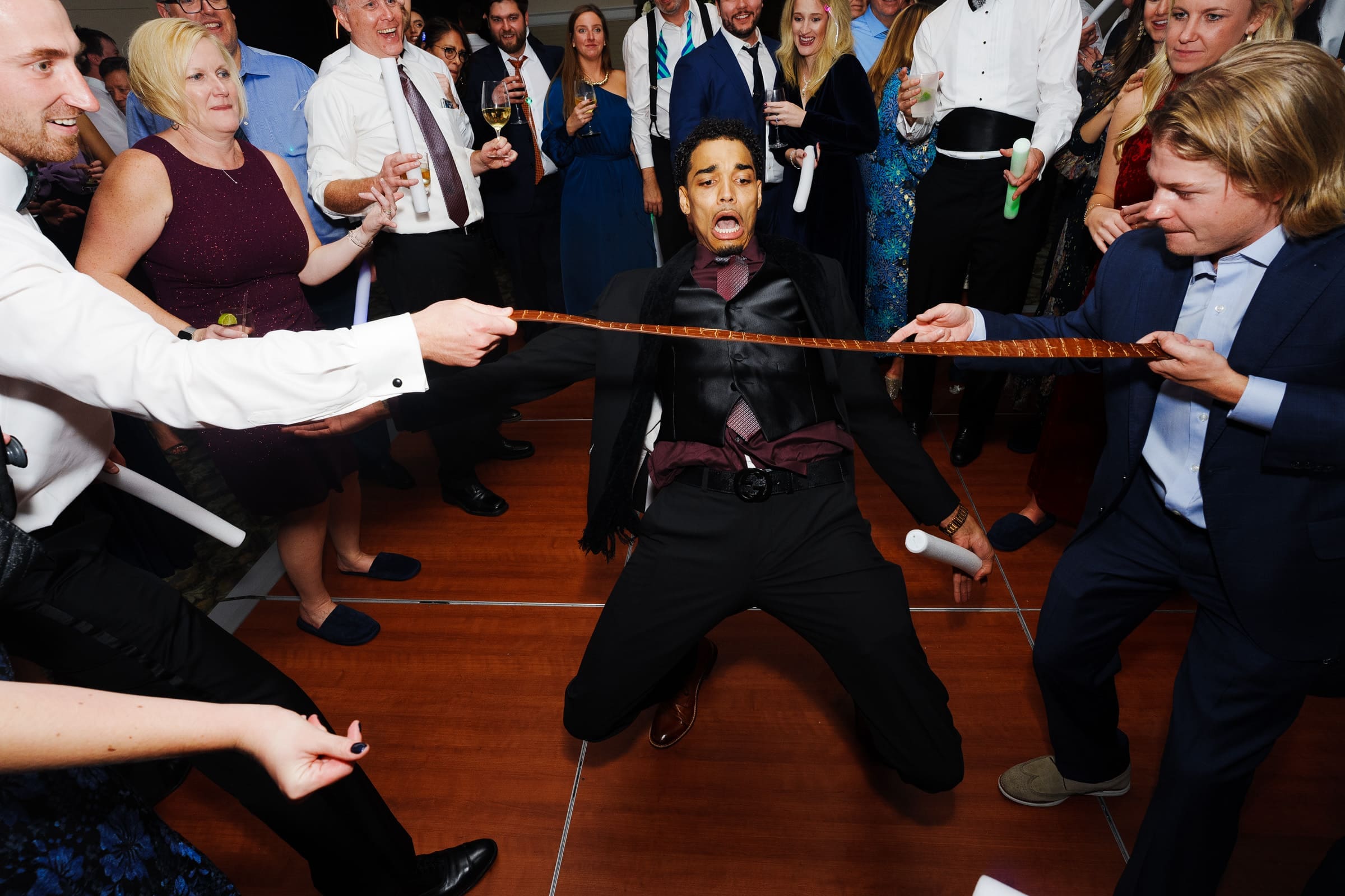 At a vibrant gathering reminiscent of Pinehurst Resort, a man in formal attire energetically does a limbo under a tie, surrounded by well-dressed people cheering him on. The group is joyful and celebratory, with colorful lights enhancing the festive atmosphere.