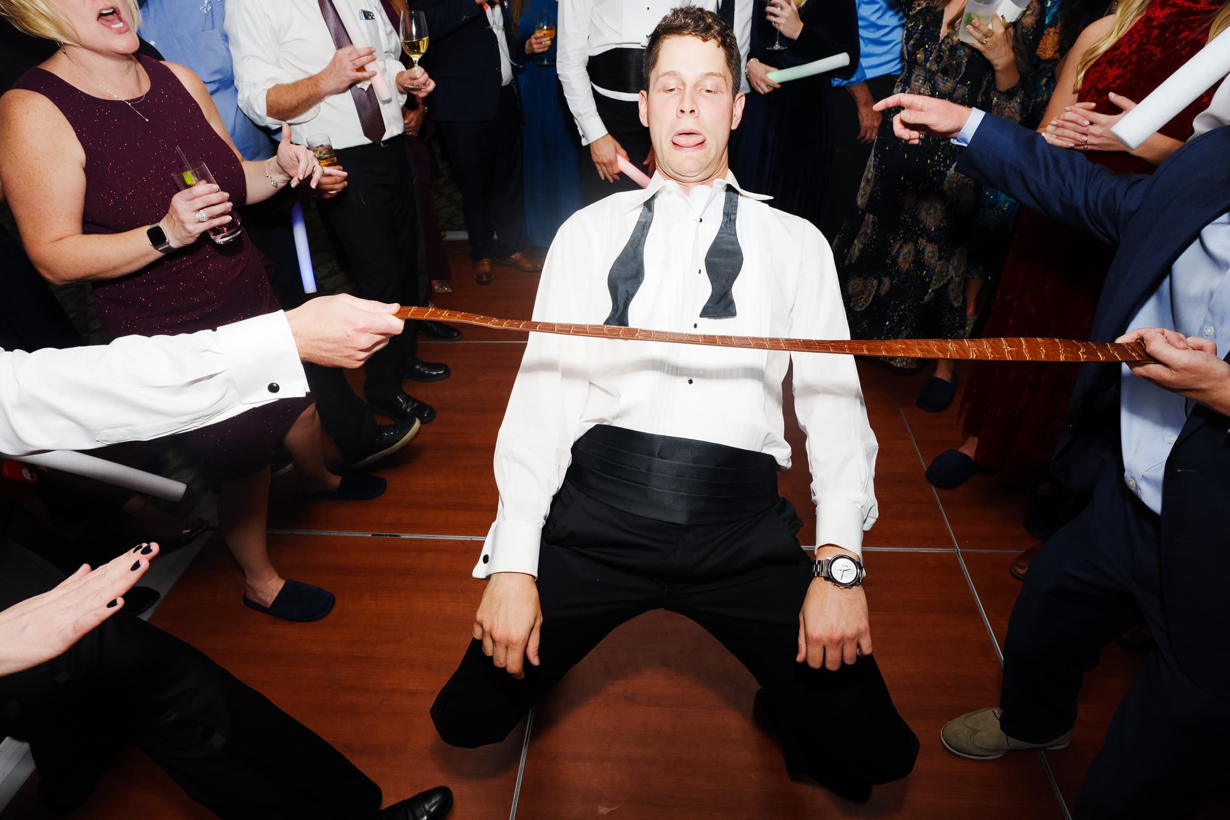 A man in a white shirt and untied black bow tie bends backward at a lively party in the Pinehurst Resort. People cheer him on, drinks in hand, as one holds the limbo stick. The festive scene mirrors the vibrant spirit often seen during celebrations near the village chapel.