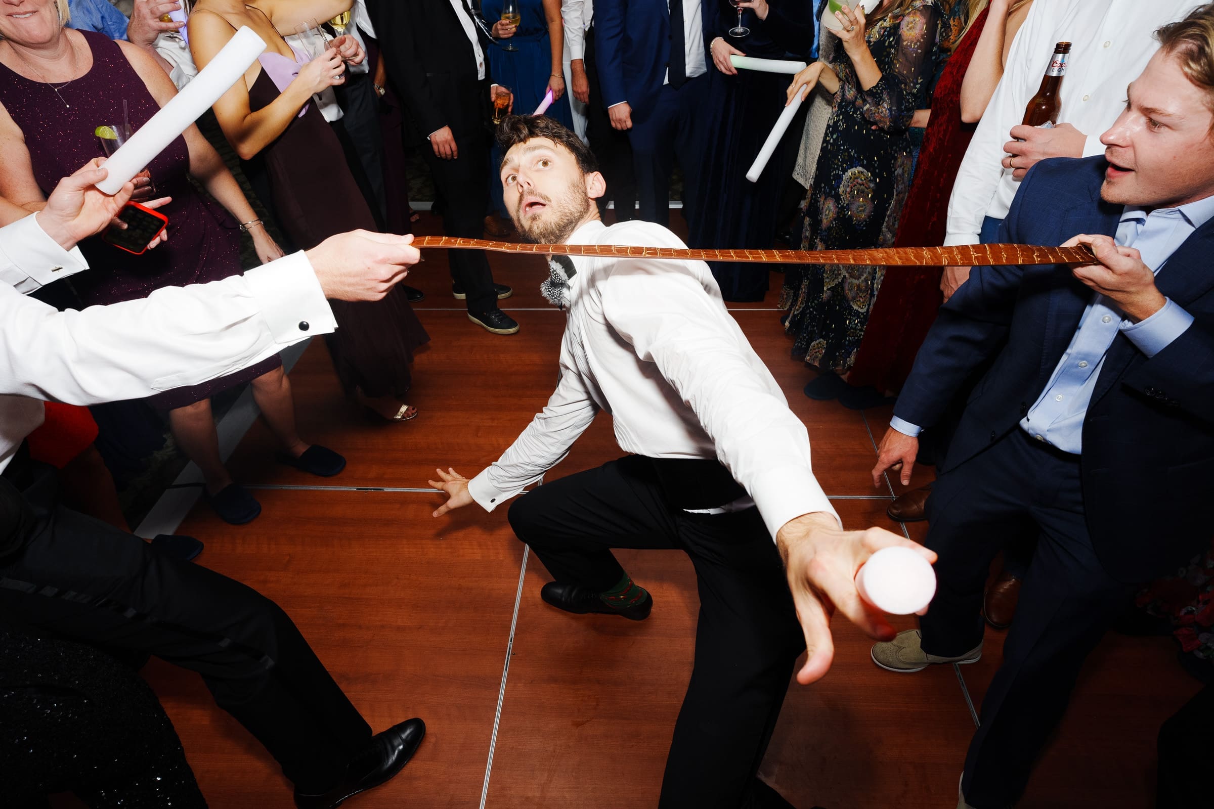 A group of people are enjoying a lively dance party at the Pinehurst Resort. A man in a white shirt and black pants bends backward to limbo under a wooden stick held by two others. Guests around him are watching and laughing, holding drinks and light sticks.