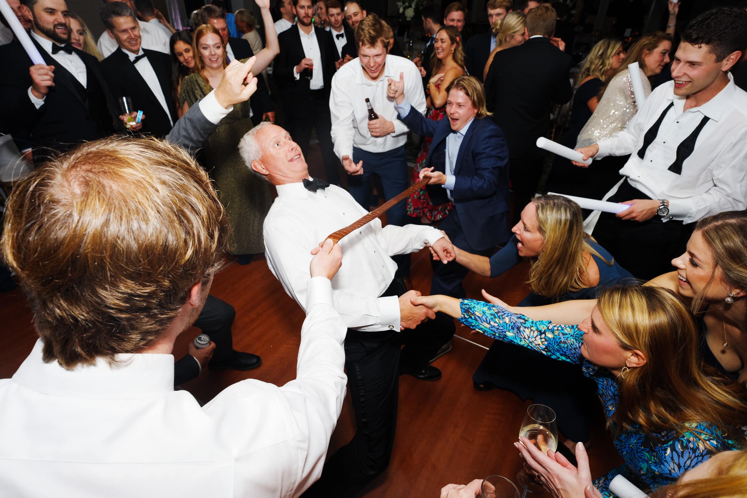 A lively wedding reception unfolds at the charming village chapel, with guests dancing and laughing energetically. A man in a white shirt playfully leans back into a limbo position, as others cheer and revel in the joyous celebration.