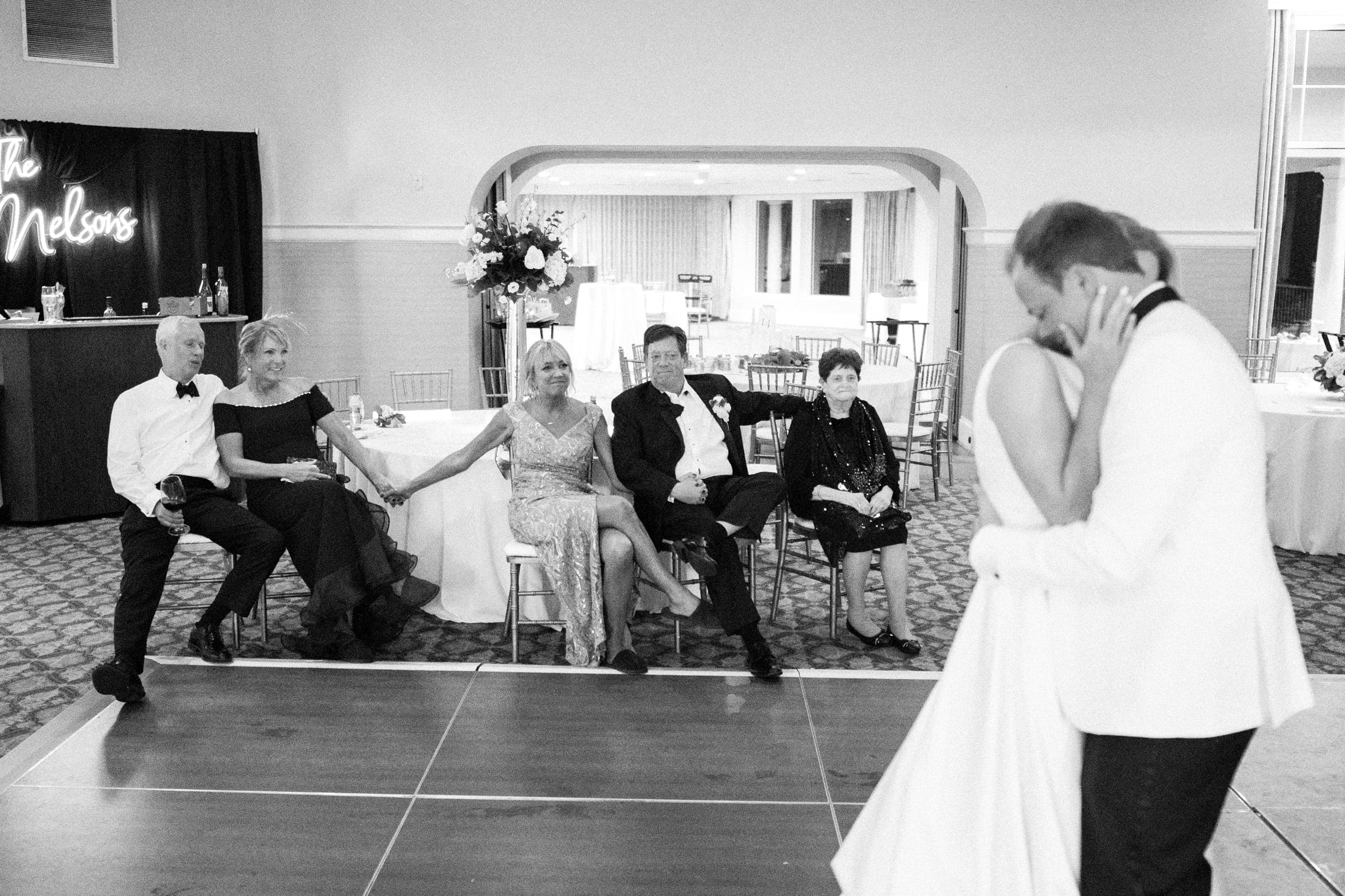 In a black and white photo, a bride and groom share a dance on the wooden floor of Pinehurst Resort. In the background, five seated guests watch as a decorative sign reading "The Nelsons" graces the left side, capturing an intimate moment of joy.