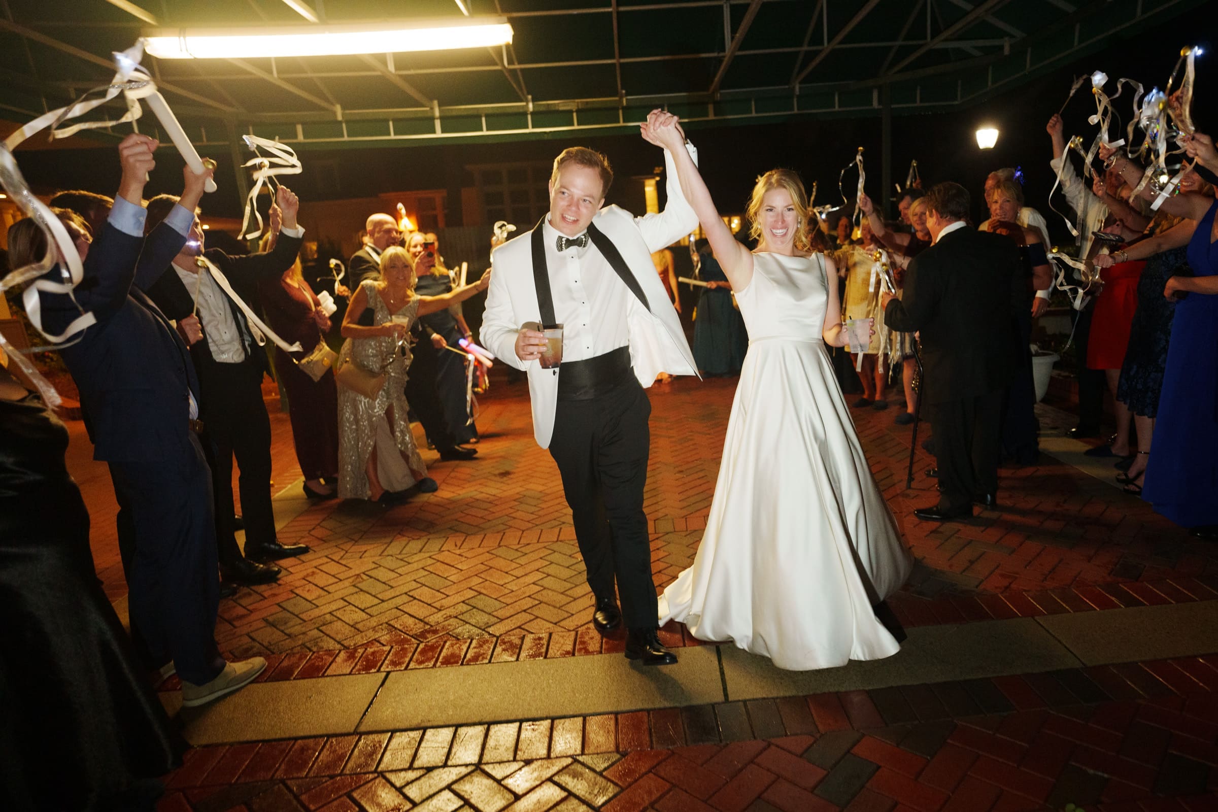 A newlywed couple walks hand in hand through a group of cheering guests outside the charming village chapel. The groom wears a white tuxedo jacket, and the bride is in a white gown. Guests wave ribbons in celebration at this joyful and festive Pinehurst Resort evening.