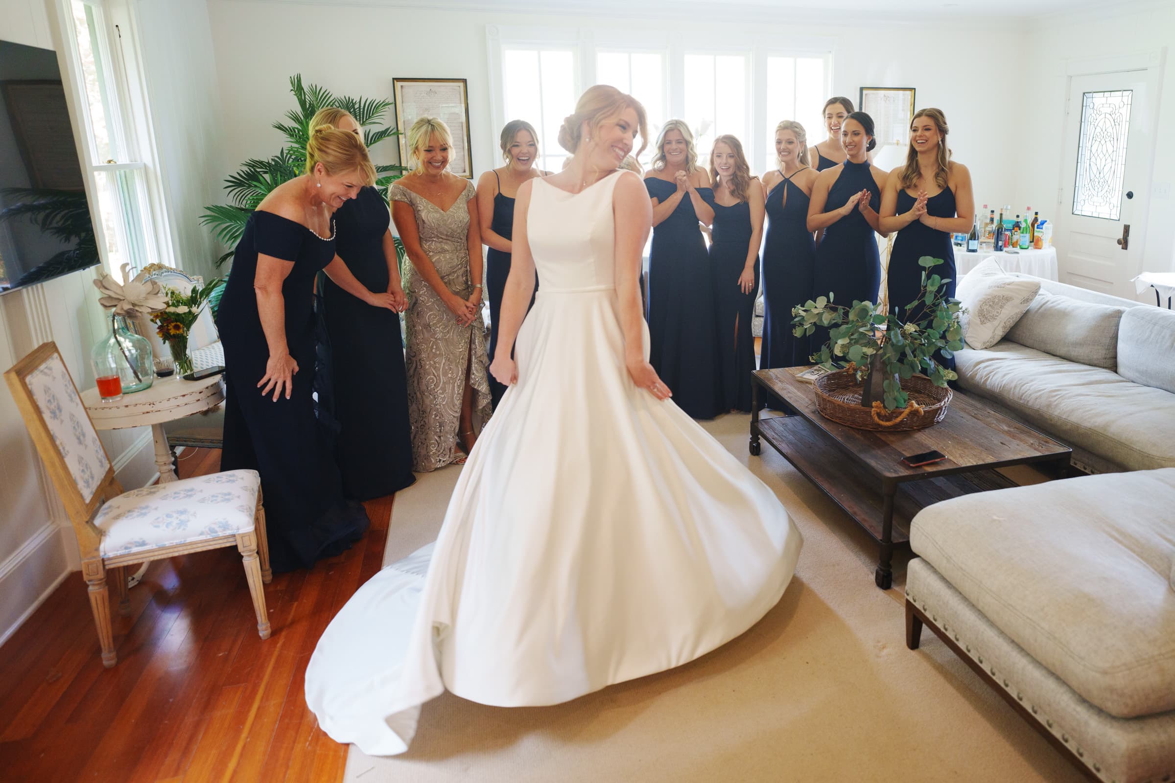 A bride in a white gown twirls joyfully in a living room, reminiscent of the elegant charm of a village chapel. Her bridesmaids, dressed in dark navy and one in silver-gray, stand behind her, clapping and smiling. The room has wooden floors, a beige sofa, and a table with decorations.