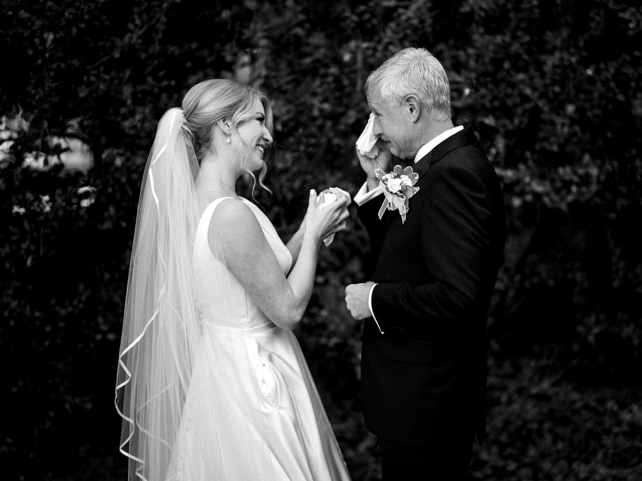 A bride and groom stand outdoors at the picturesque Pinehurst Resort, smiling and looking at each other. The bride wears a flowing gown and veil, holding a tissue, while the groom, in a suit with a boutonniere, wipes his eye. The backdrop features blurred greenery reminiscent of a village chapel setting.