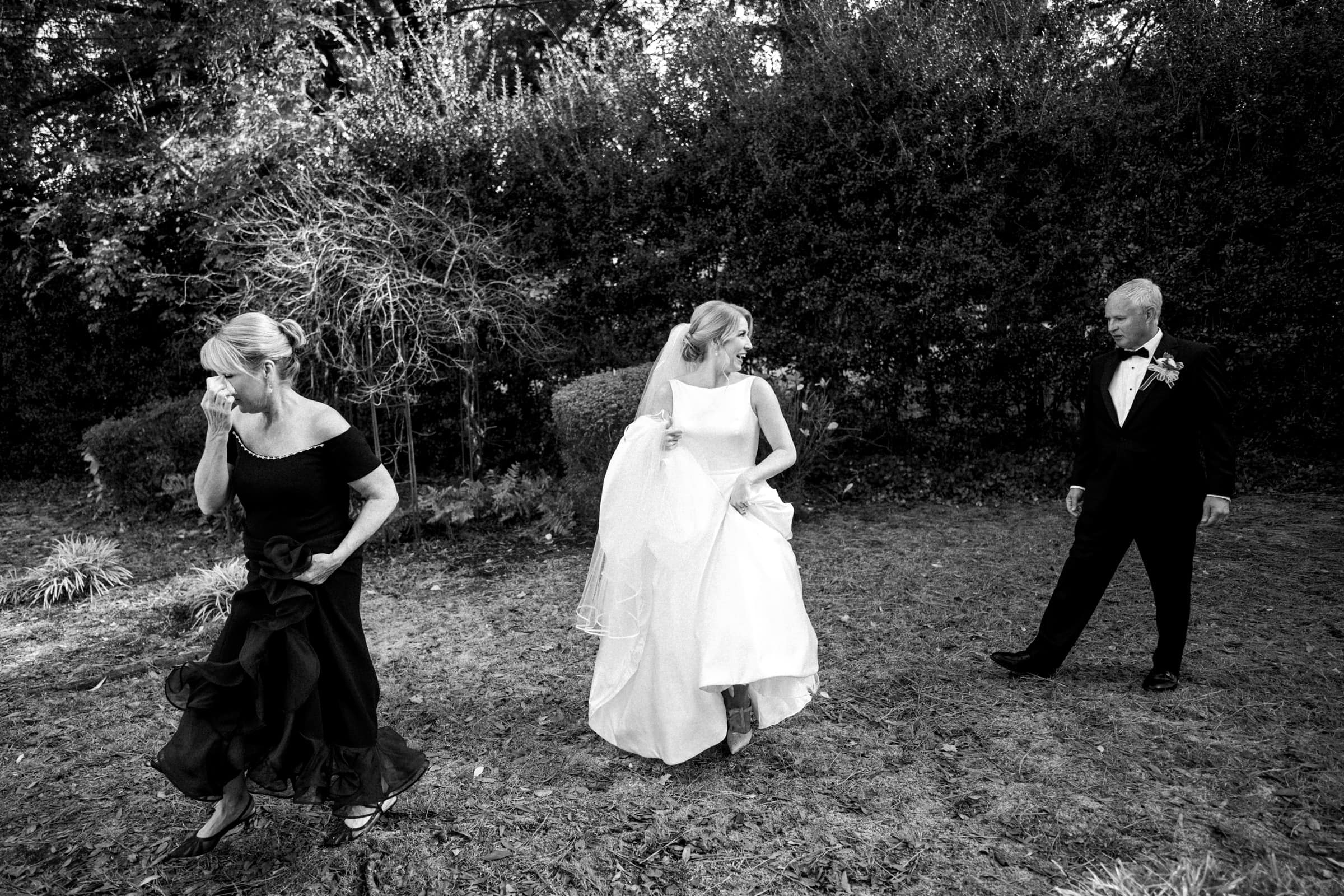 A bride in a white gown walks across a grassy area near the village chapel, holding her dress. A woman in a dark gown walks ahead of her, and a man in a tuxedo follows behind. The scene at Pinehurst Resort is set outdoors with trees and bushes in the background.