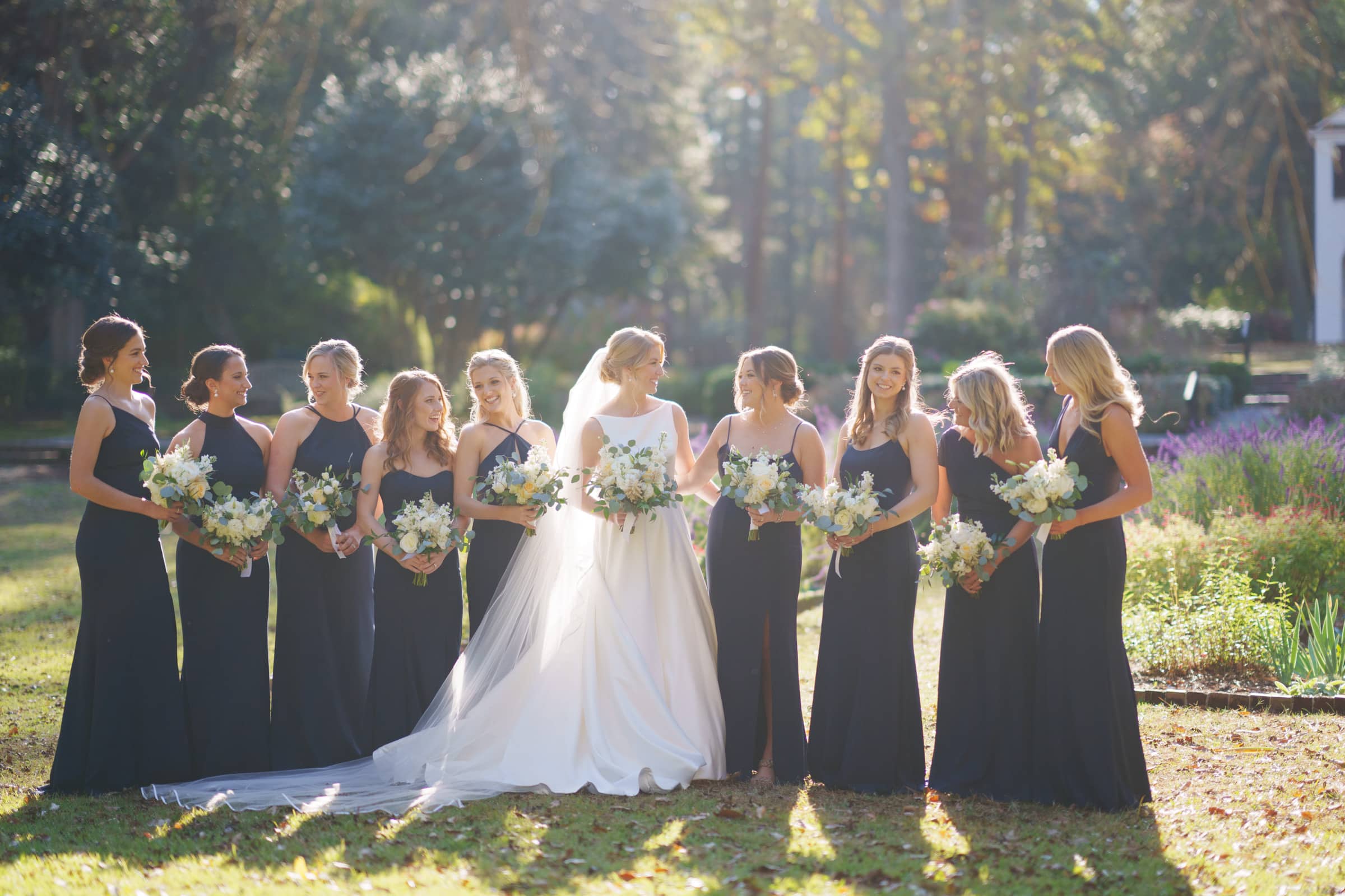 A bride stands with her bridesmaids in a sunlit garden at the charming village chapel. The bride is in a white gown, while the bridesmaids wear matching dark dresses. Each holds a bouquet of white and green flowers. Sunlight filters through the trees, creating a warm, serene atmosphere.