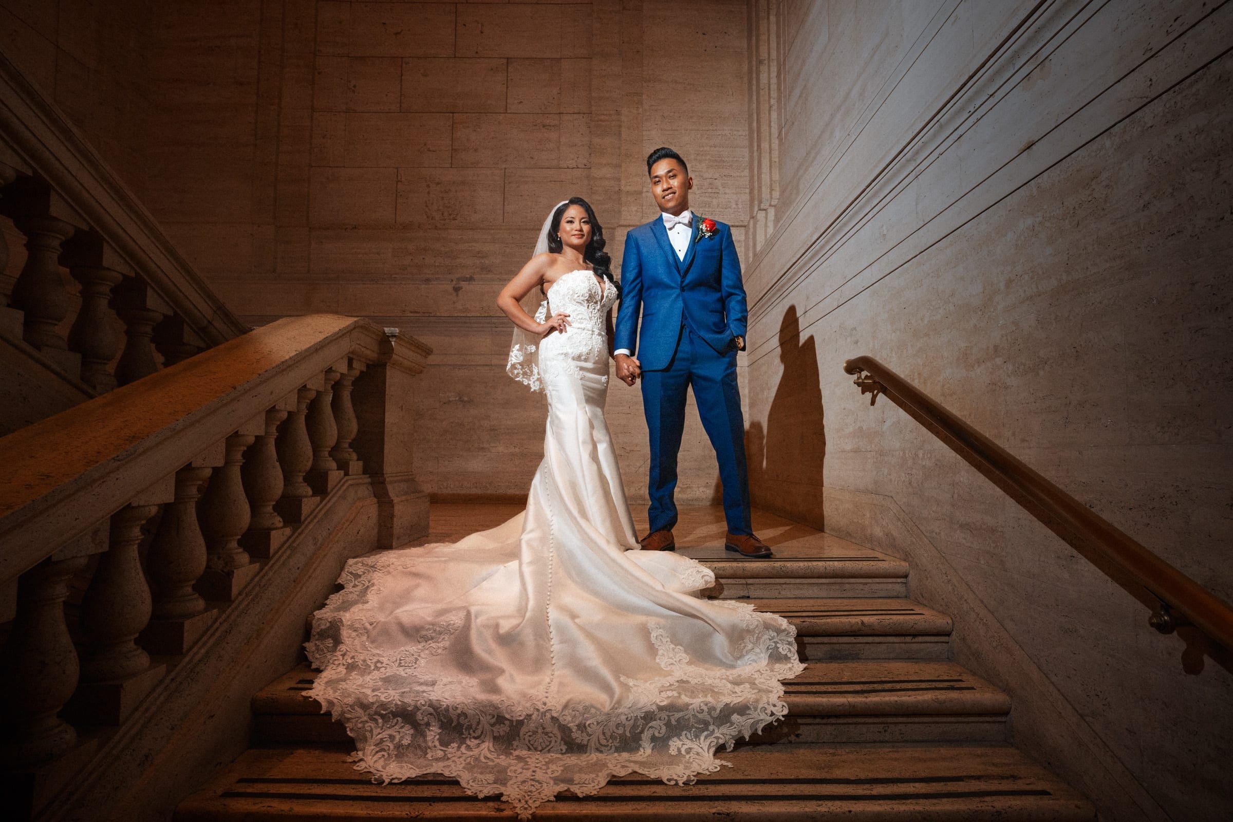 A bride and groom stand on a staircase in formal wedding attire, captured perfectly by downtown Chicago wedding photography. The bride wears a long, elegant white gown with a lace train, while the groom is in a blue suit with a red boutonniere. They hold hands, posing for a photo on the stone steps.