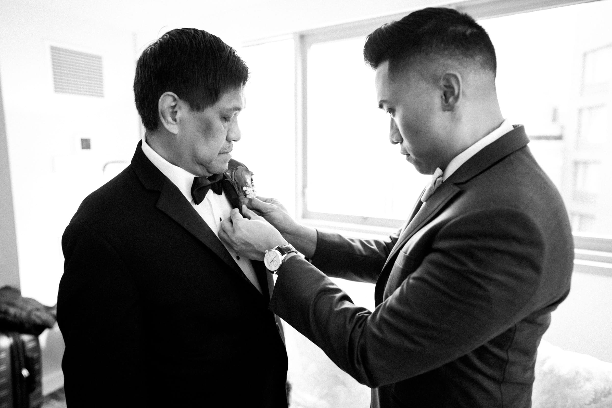 downtown chicago wedding photography - rena & von In a sunlit room with a view of downtown Chicago's skyline, a young man assists an older gentleman in perfecting his bow tie. Both focused and elegantly suited, they share a moment captured with the essence of wedding photography, as natural light floods through the window.