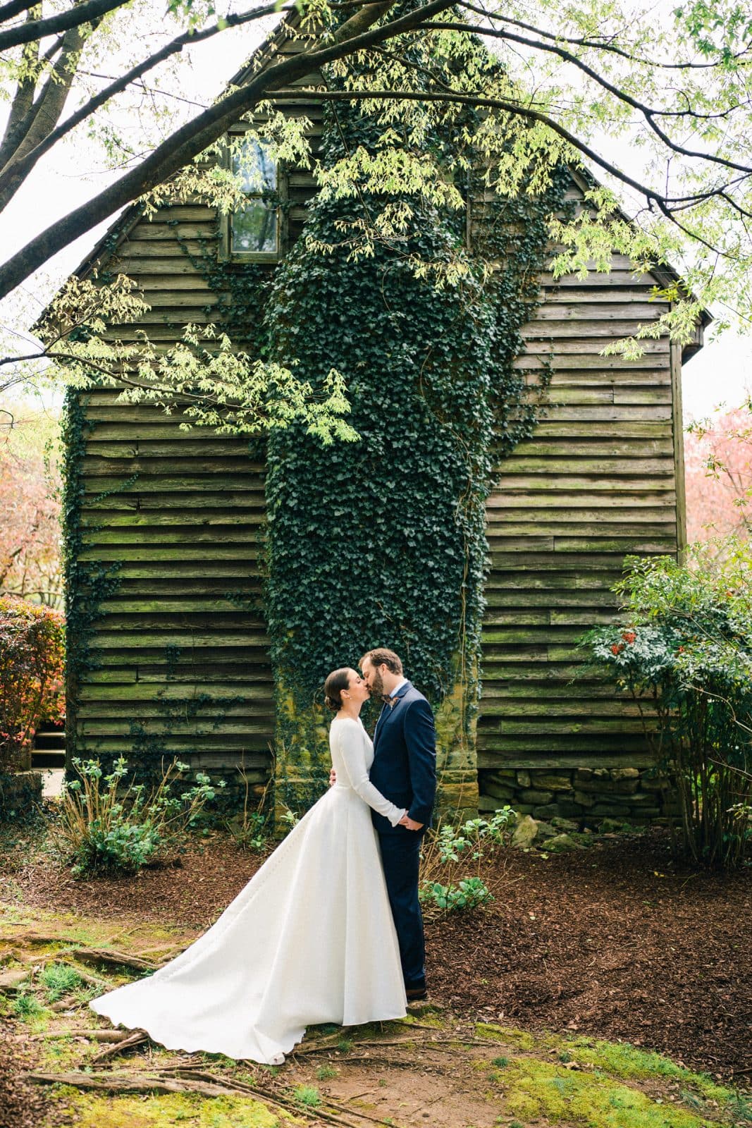 A bride and groom stand holding hands and kissing in front of a rustic wooden building covered in ivy at their Sutherland Estate wedding. The bride wears a white dress, while the groom is in a dark suit. Trees with green leaves arch overhead, creating a natural, serene setting.
