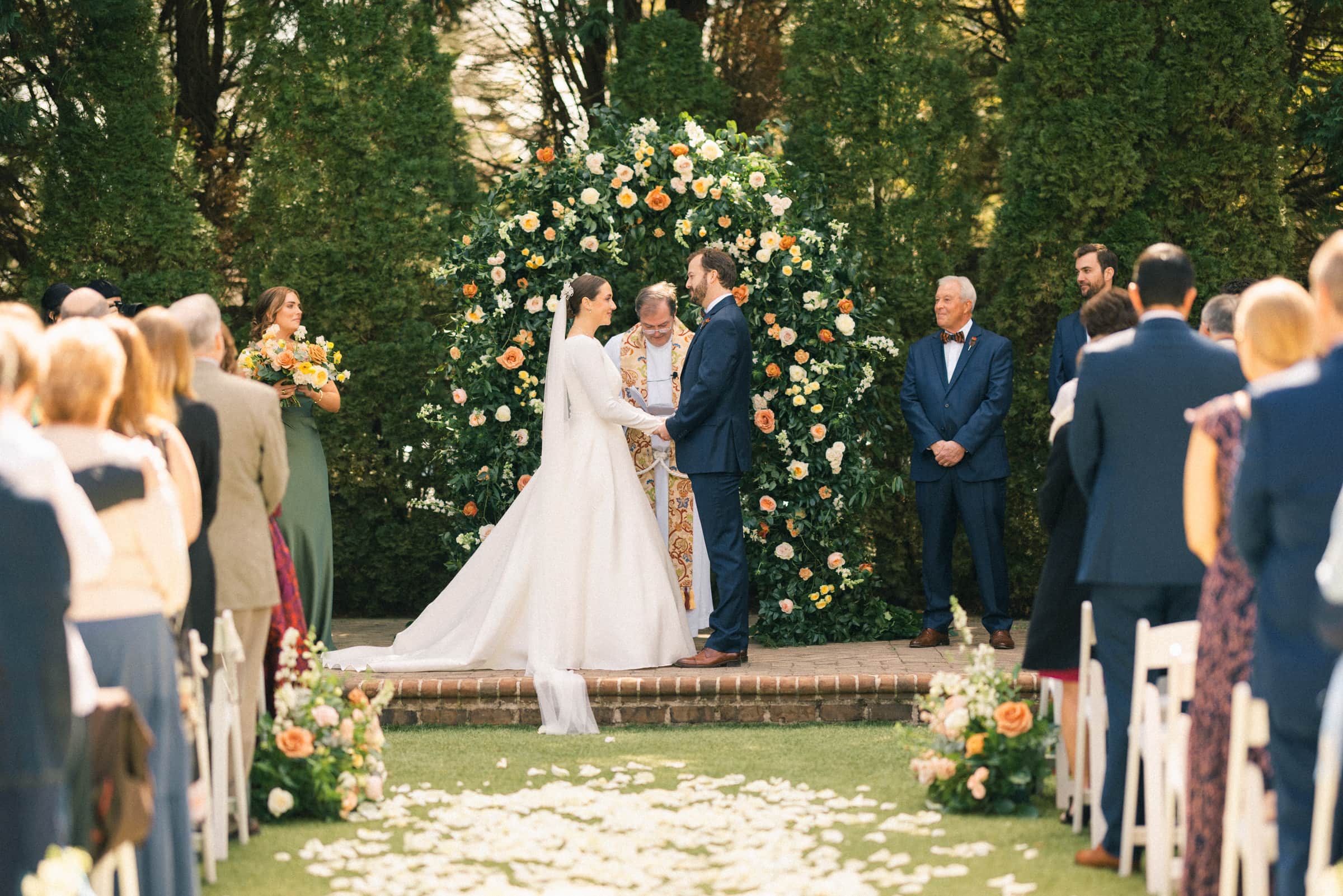 A Sutherland Estate wedding unfolds as a couple exchanges vows beneath a stunning floral arch. Guests sit on either side of a petal-lined aisle, while the officiant stands poised between them, and attendants are elegantly positioned on both sides, enhancing the enchanting outdoor ceremony.